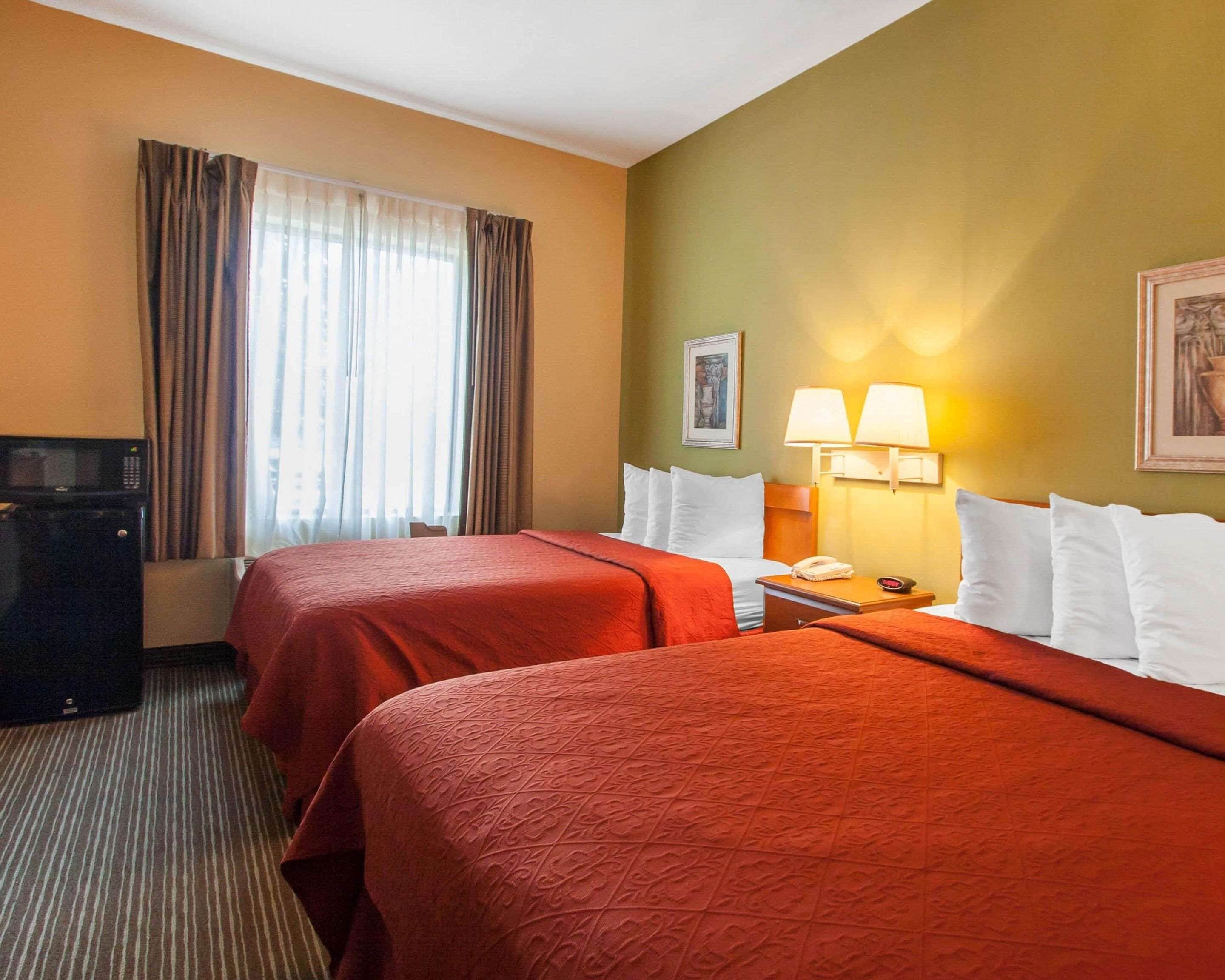 A hotel room with two beds, a desk, a lamp, a microwave, and a painting on the wall.