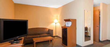 A hotel room with a flat-screen TV, a sofa, a small table, a lamp, a microwave, and a door leading to another room.