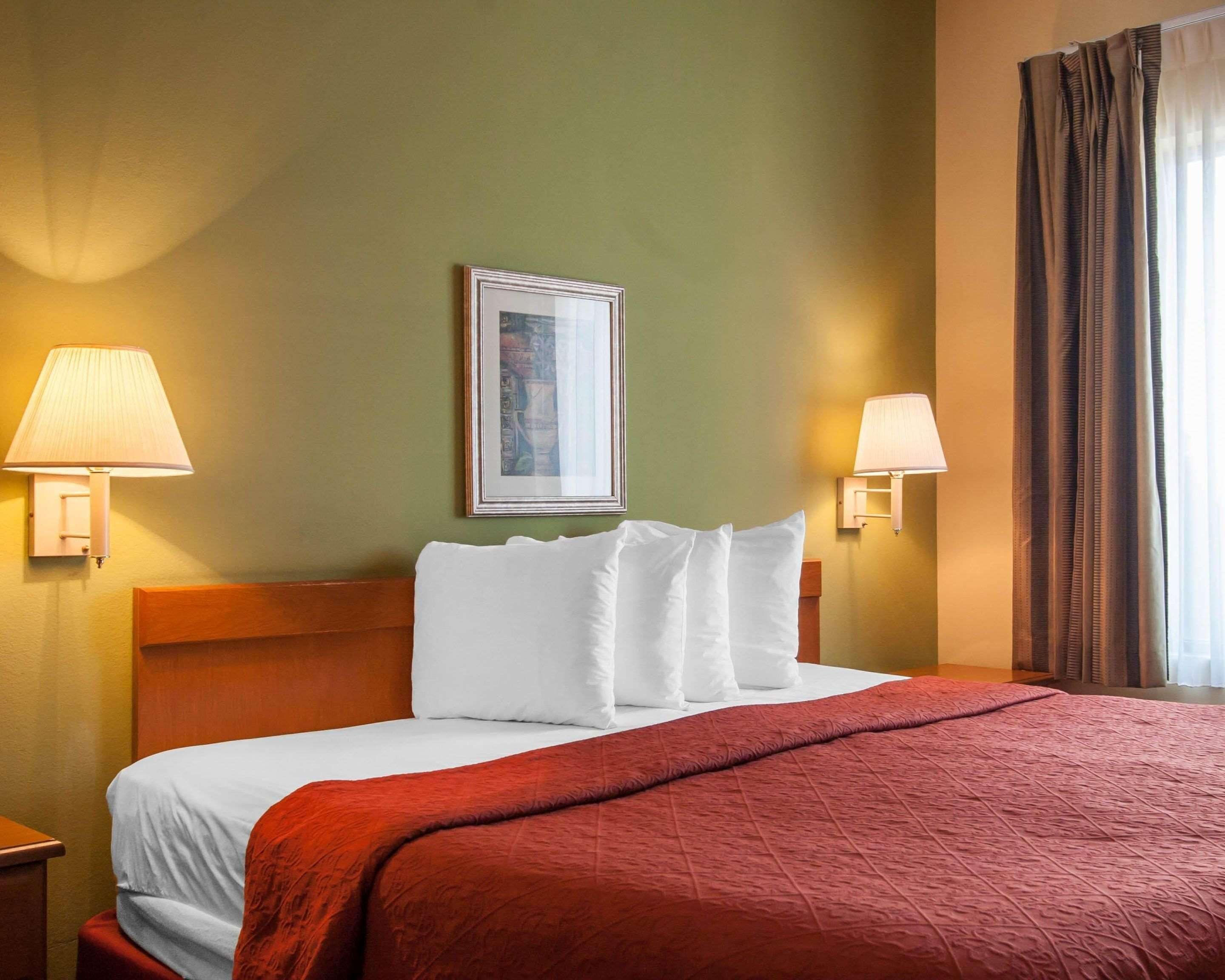 Ein Hotelzimmer mit einem Bett, zwei Wandlampen, einem gerahmten Bild und einem Fenster mit Vorhängen.
