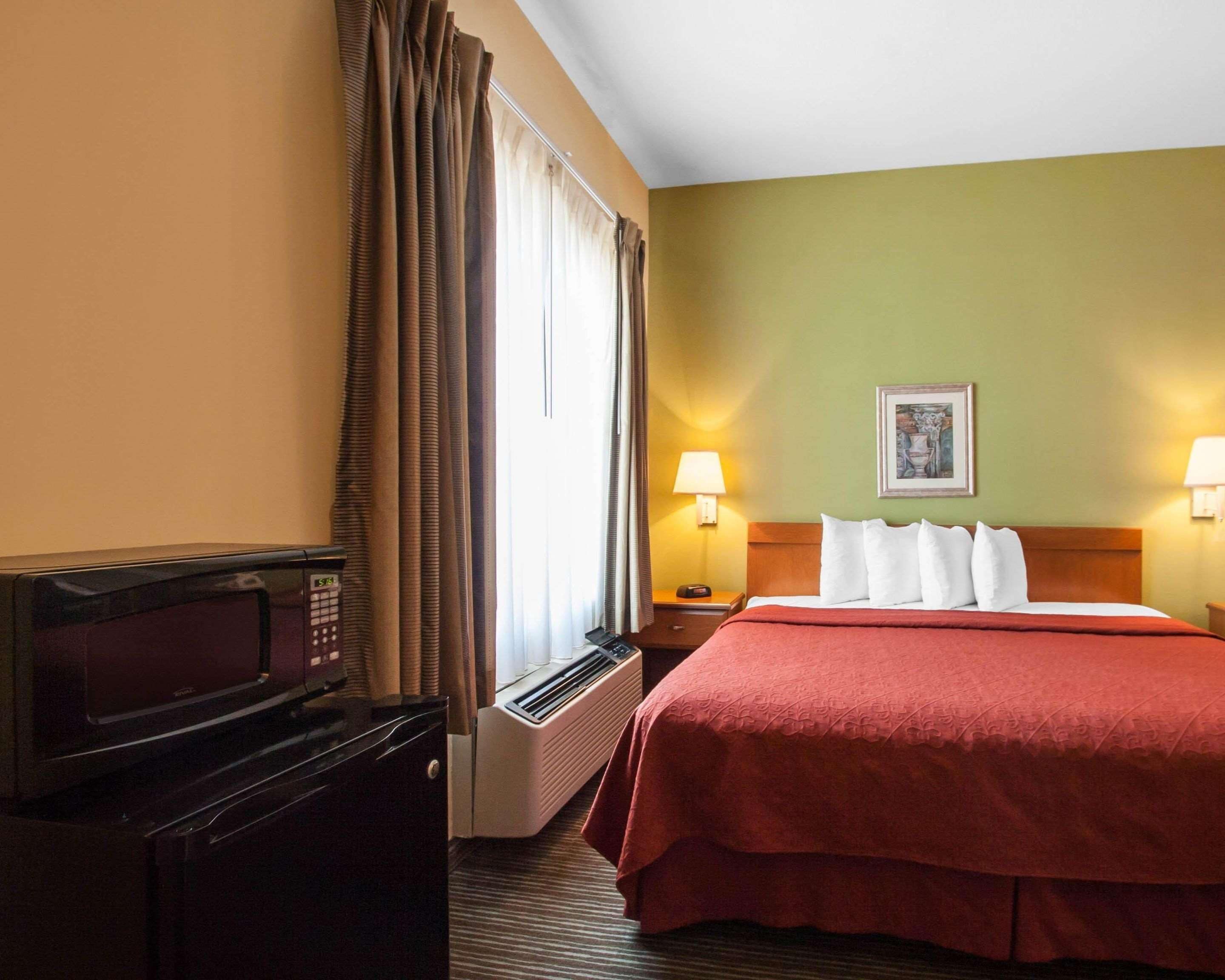 A hotel room with a bed, two bedside lamps, a heater, a microwave, and a window with curtains.