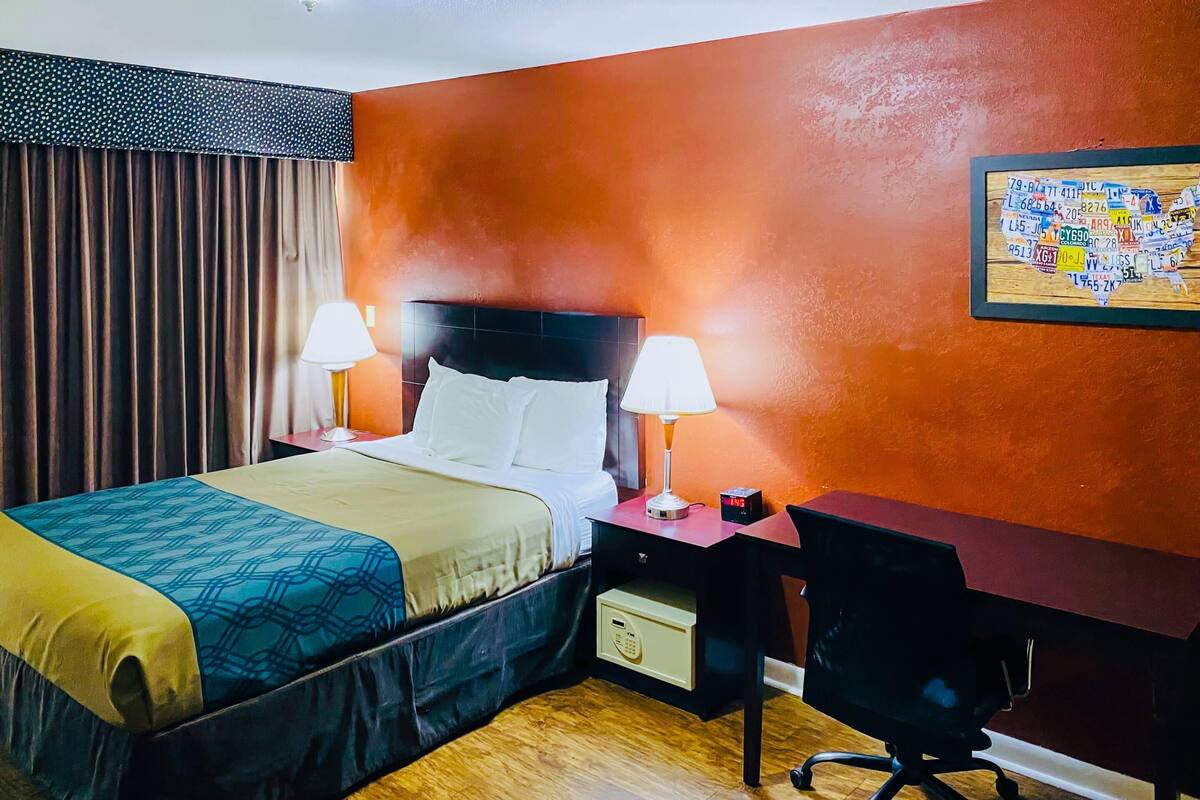 A hotel room with a bed, two bedside lamps, a desk with a chair, and a wall clock.
