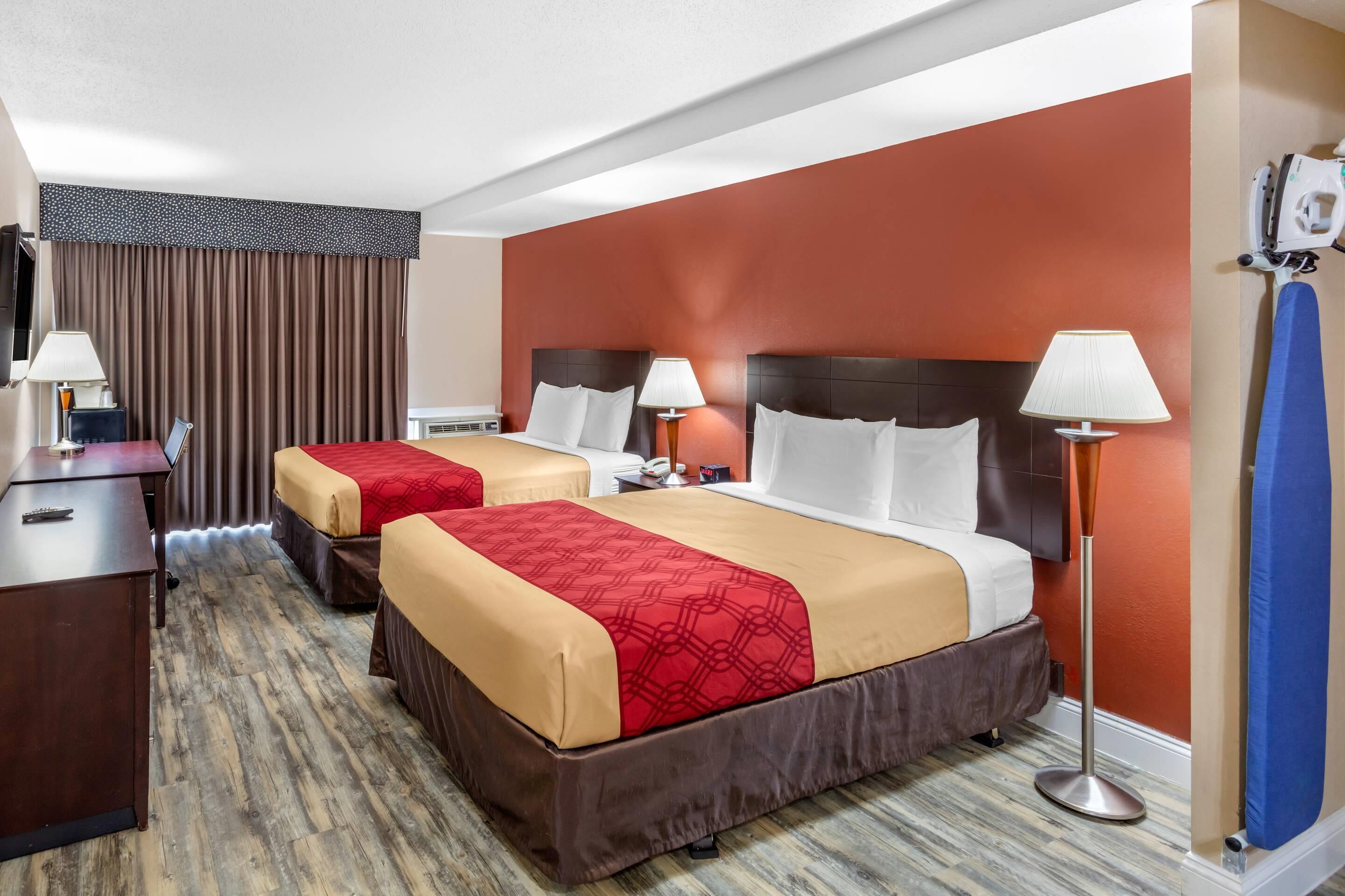 Habitación de hotel con dos camas, cada una con cubrecama rojo y beige, suelo de madera, escritorio con lámpara y tabla de planchar fijada a la pared.