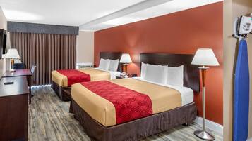 Habitación de hotel con dos camas, cada una con cubrecama rojo y beige, suelo de madera, escritorio con lámpara y tabla de planchar fijada a la pared.