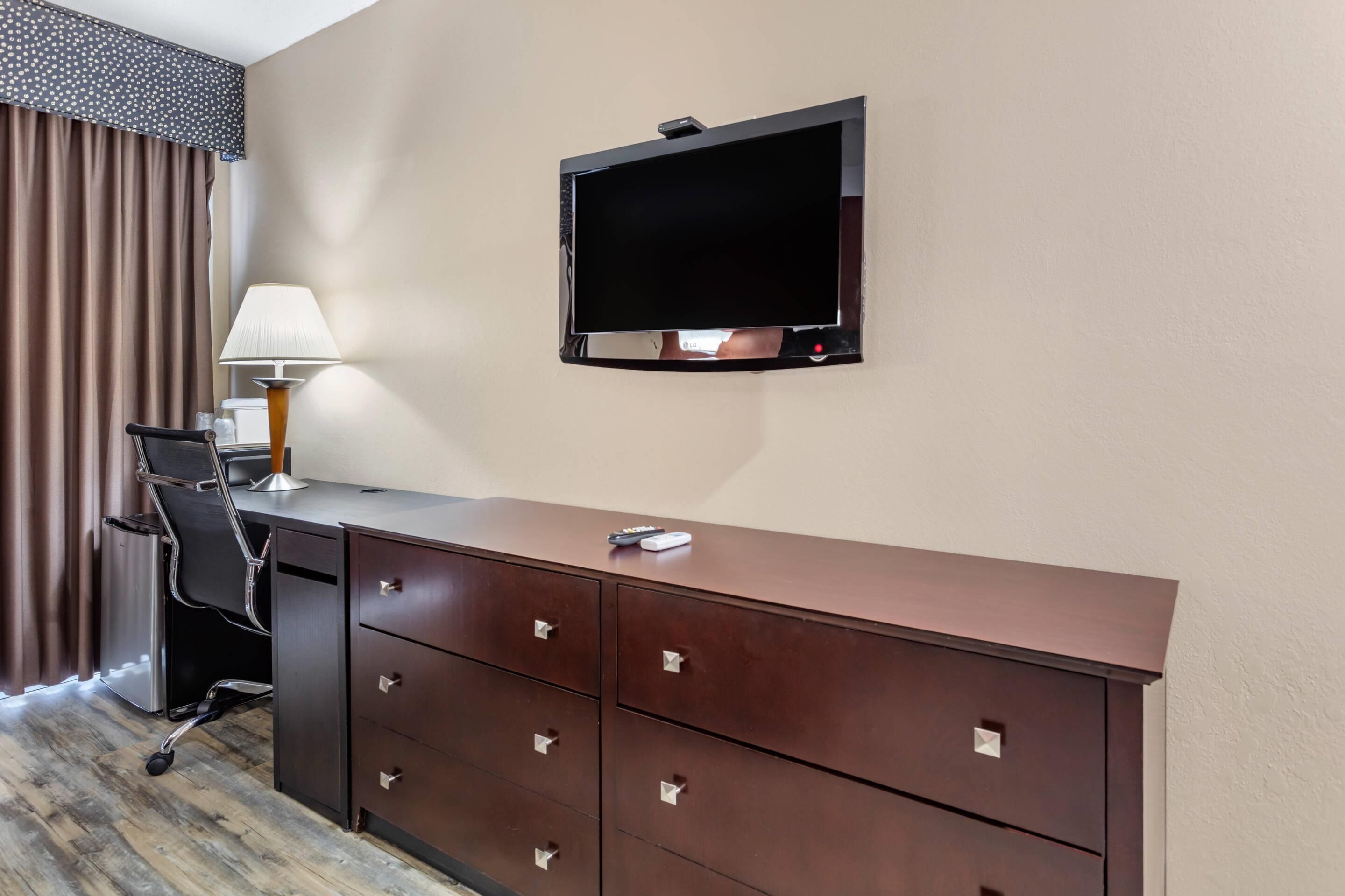 Habitación de hotel con escritorio, televisor montado en la pared, lámpara y una silla.