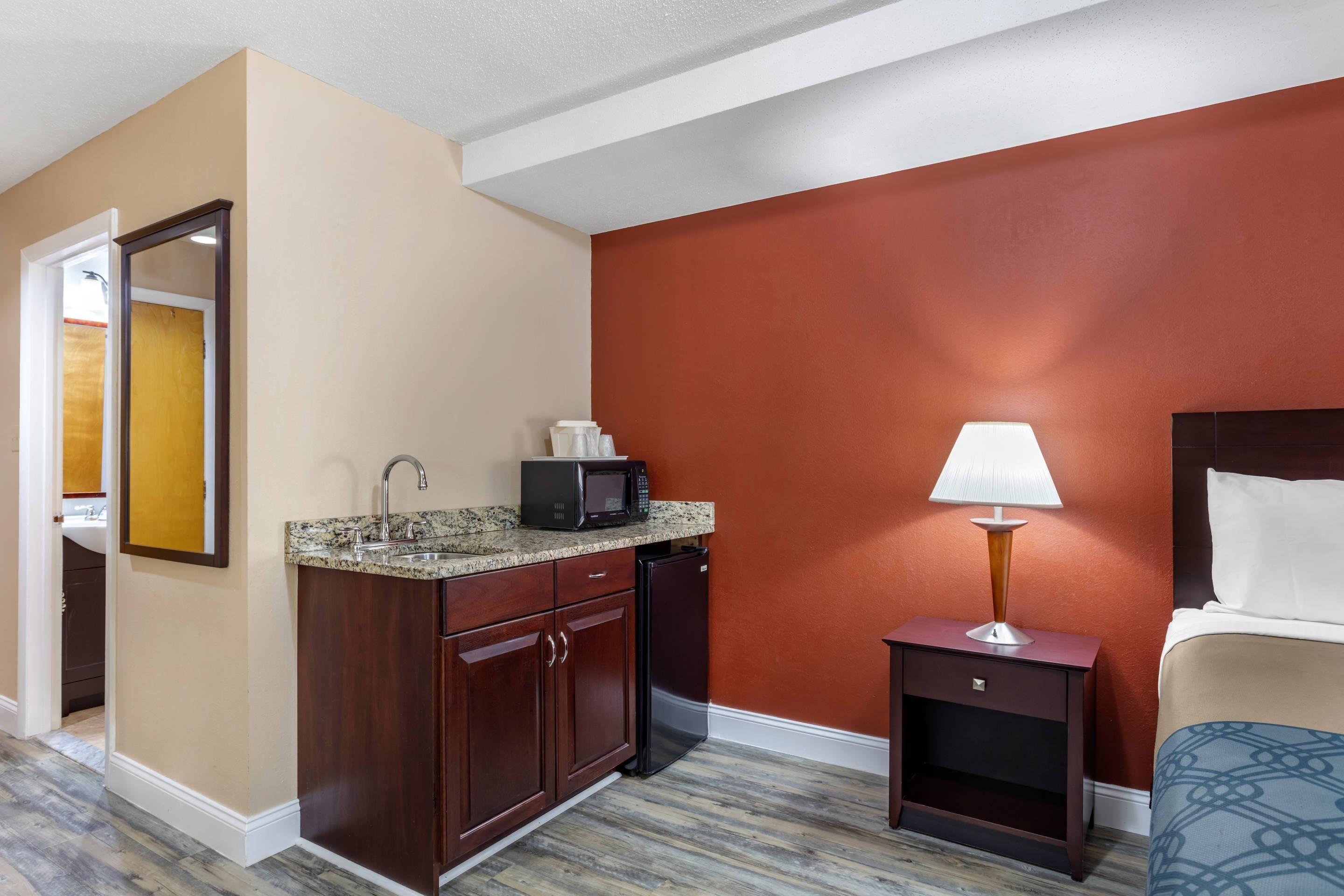 A hotel room with a kitchenette, a bed with patterned bedding, a nightstand with a lamp, and a wooden floor.