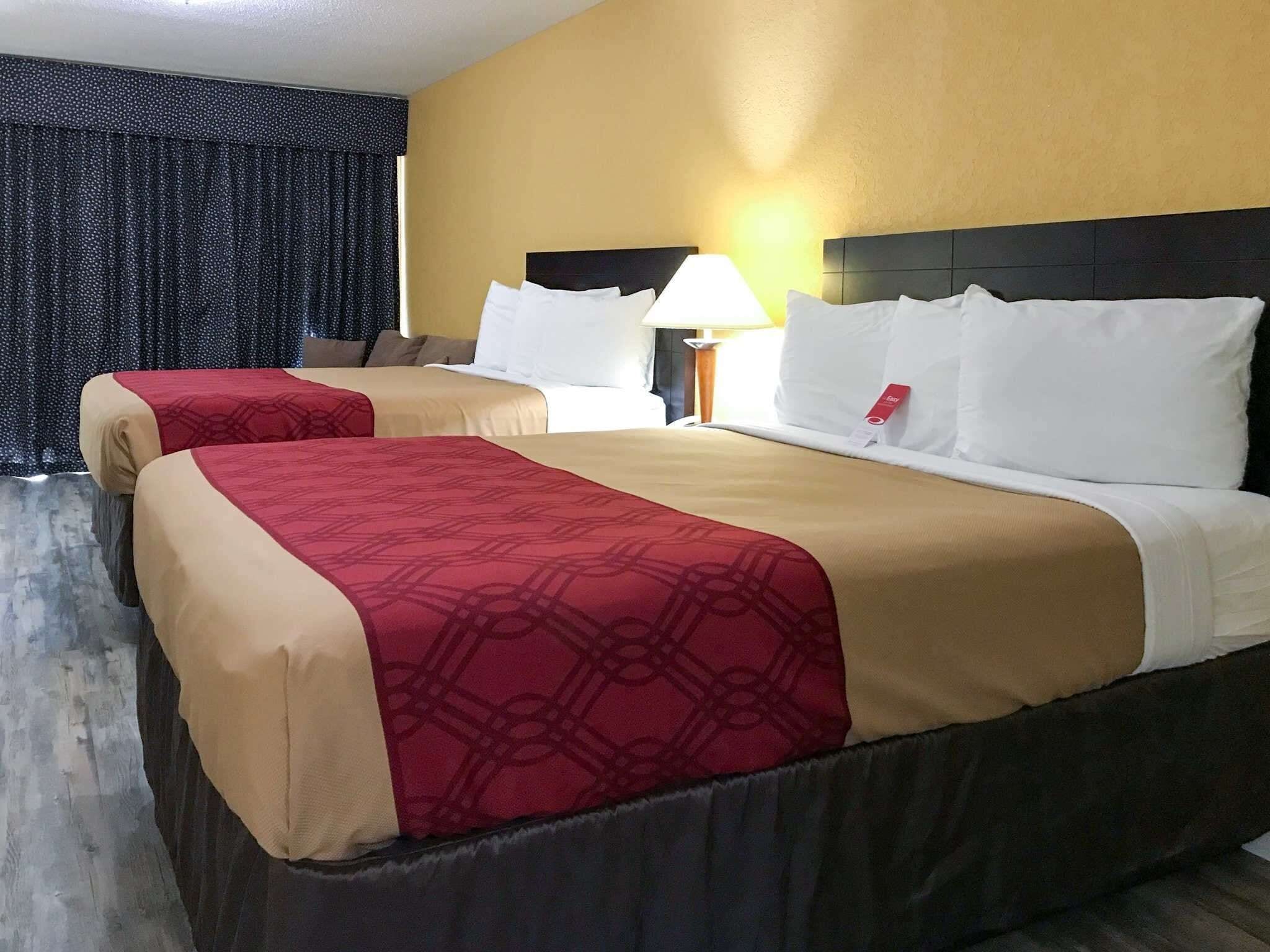 Habitación de hotel con dos camas, cada una con colcha color beige y rojo, almohadas blancas y cabecero negro. Se observa un mesita de noche con lámpara y una etiqueta roja en la almohada.