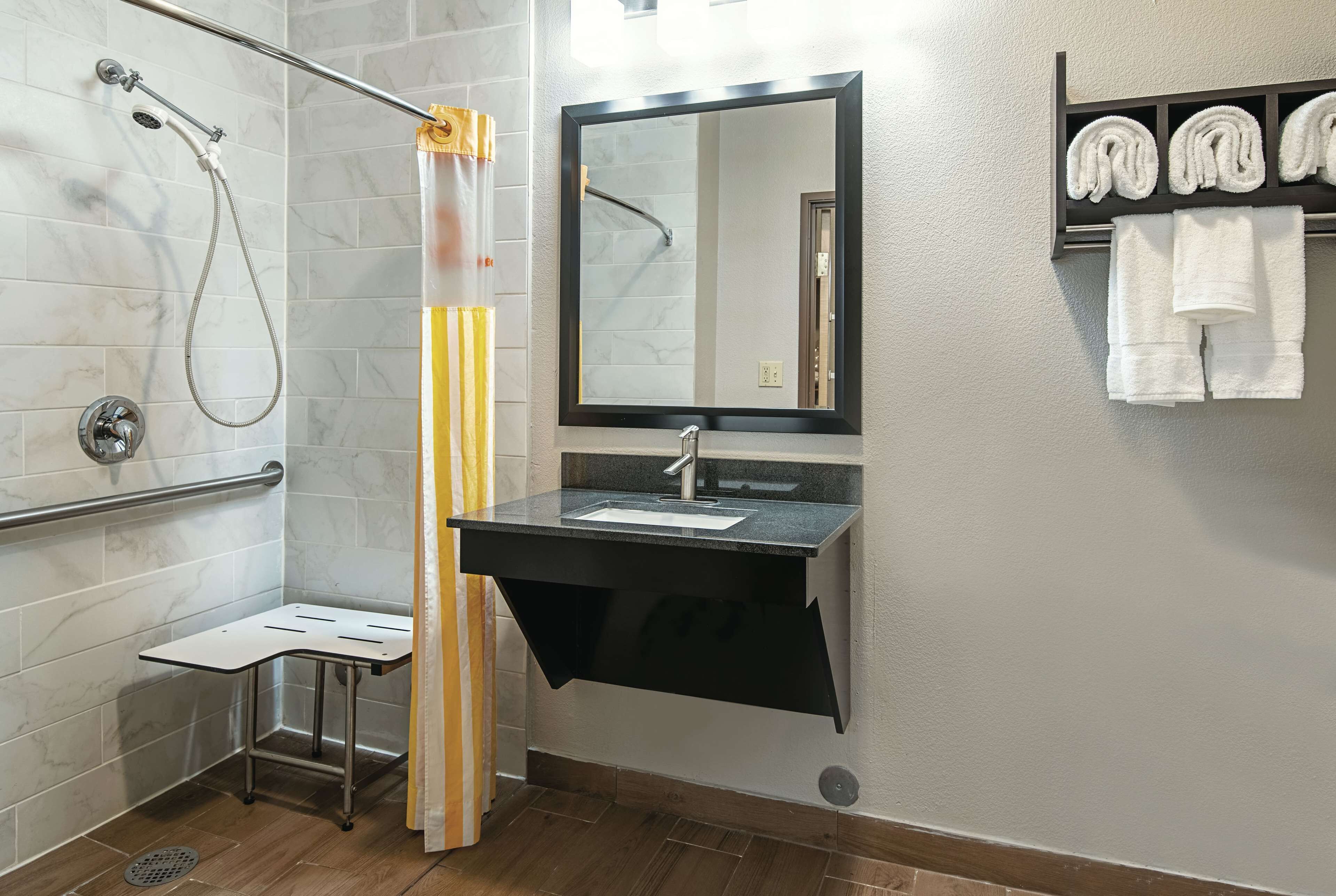 A bathroom with a shower, a sink with a mirror, a towel rack, and a small stool.
