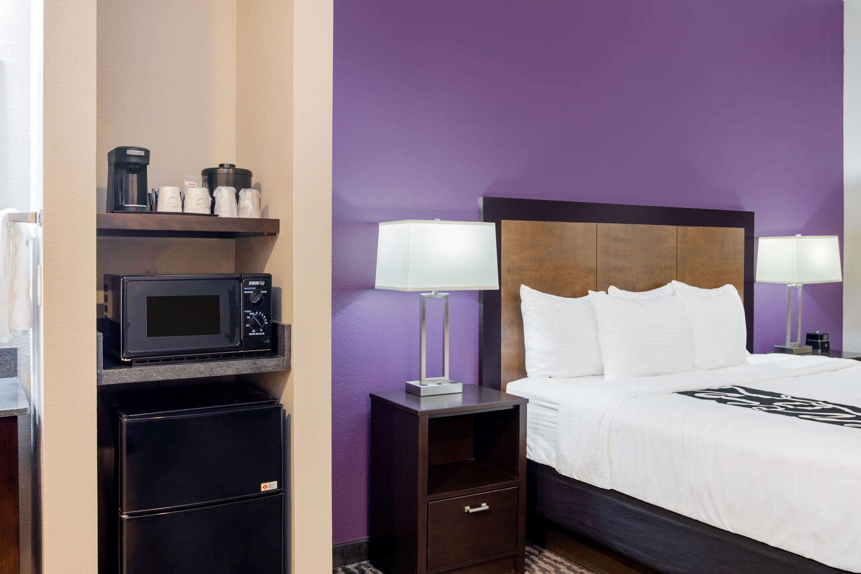 A hotel room with a bed, a nightstand with a lamp, a microwave, and a refrigerator.