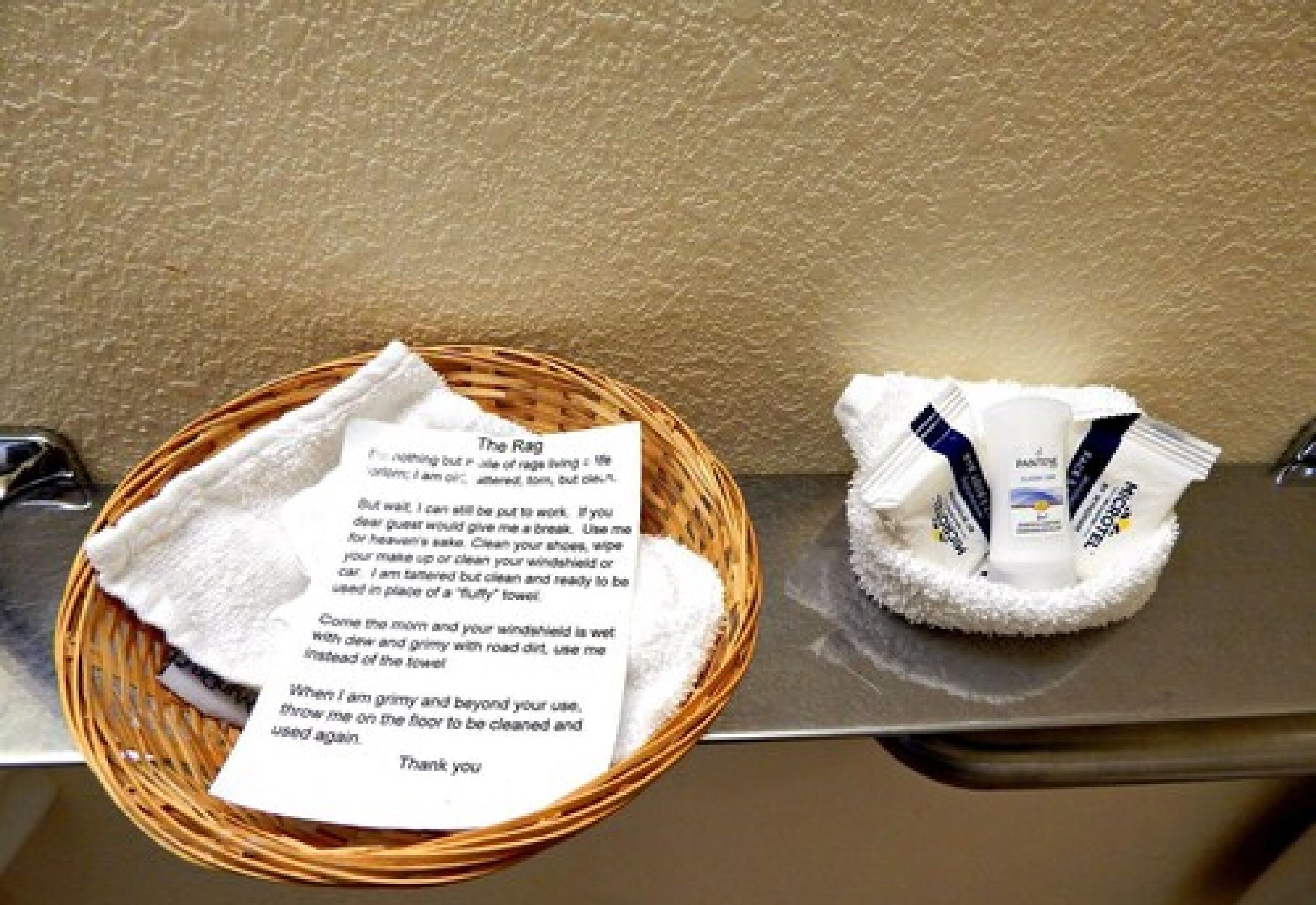 Una cesta de mimbre con una toalla doblada y una nota, y una alfombra de baño blanca sobre una superficie metálica.