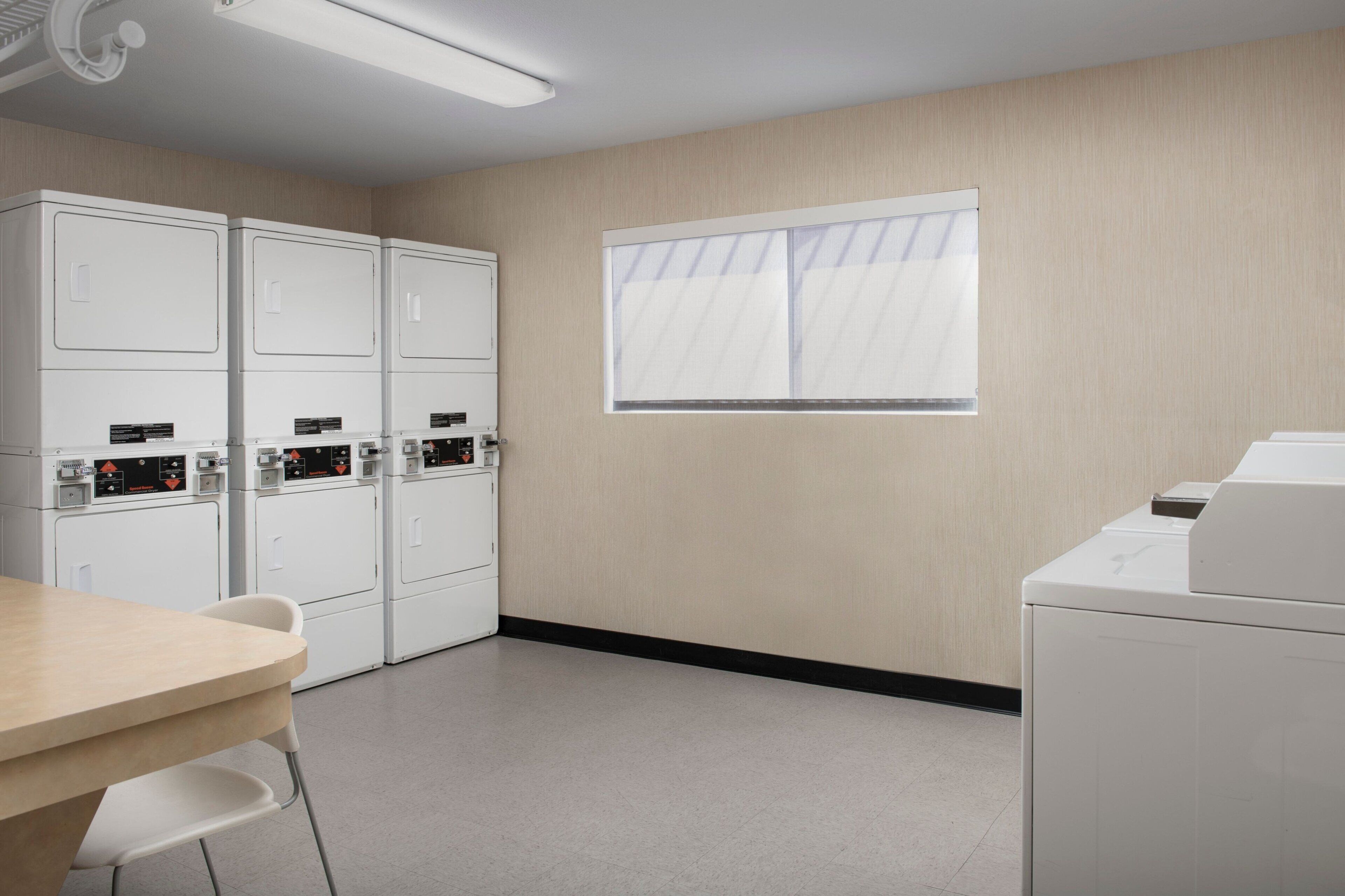 A laundry room with four white washing machines, a table with chairs, and a window with blinds.