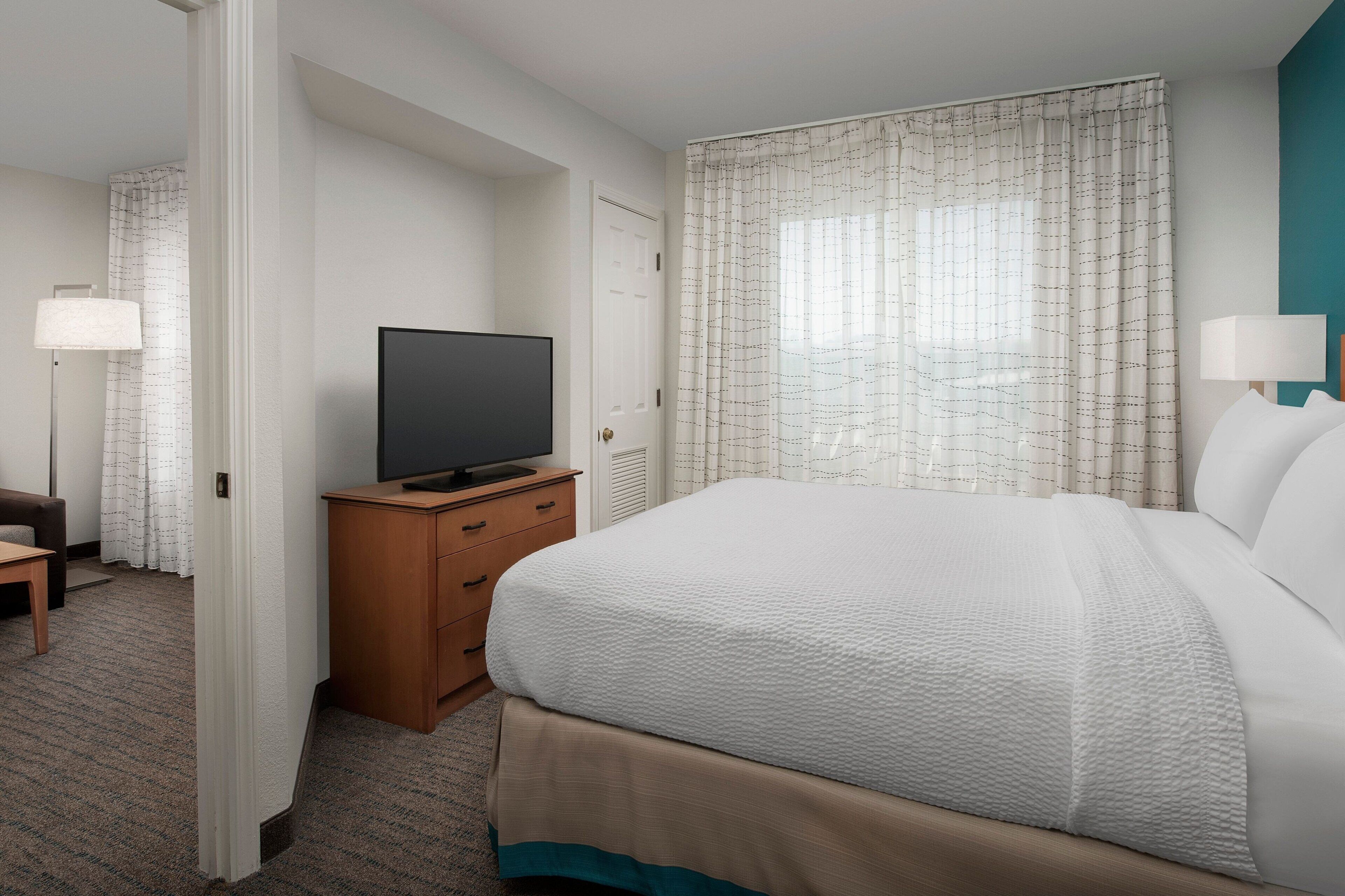 A hotel room with a large bed, a television on a wooden dresser, and a view of the outdoors through sheer curtains.