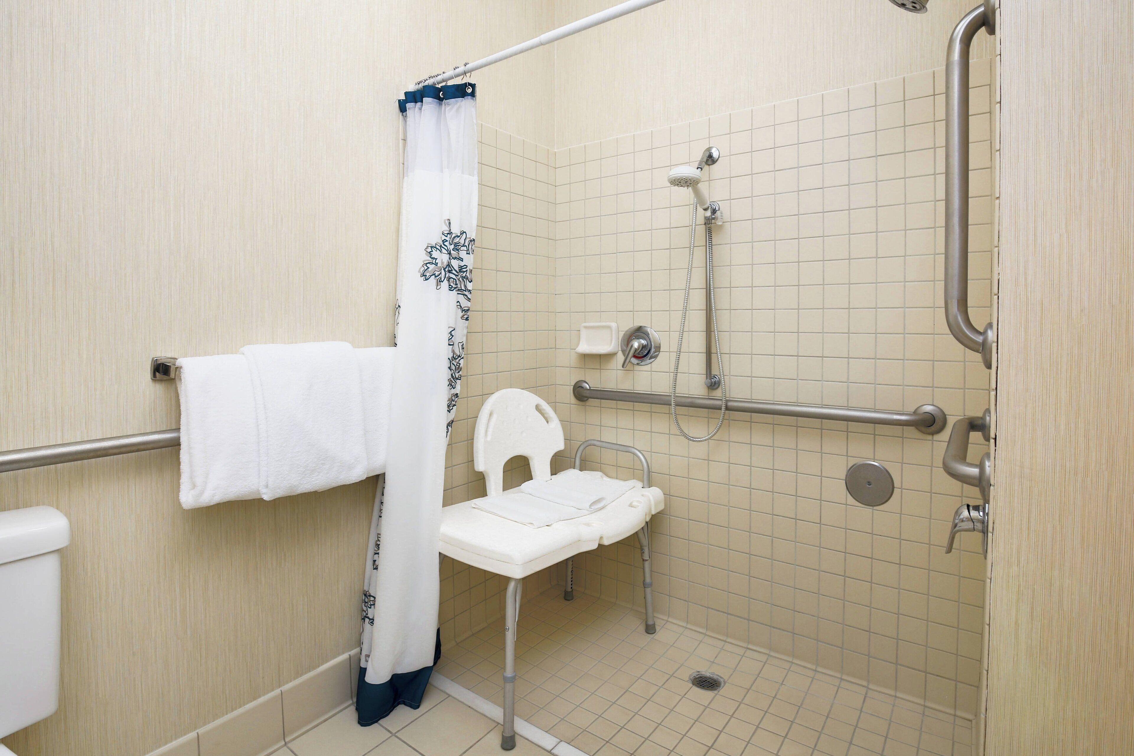 A wheelchair-accessible shower area with a grab bar, a white chair, and a towel rack.