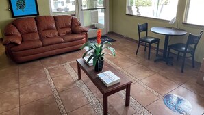 A living room with a brown leather sofa, a small table with a plant, and a window with a view of the outside. - Motel 6 Riviera Beach, FL (West Palm Beach)