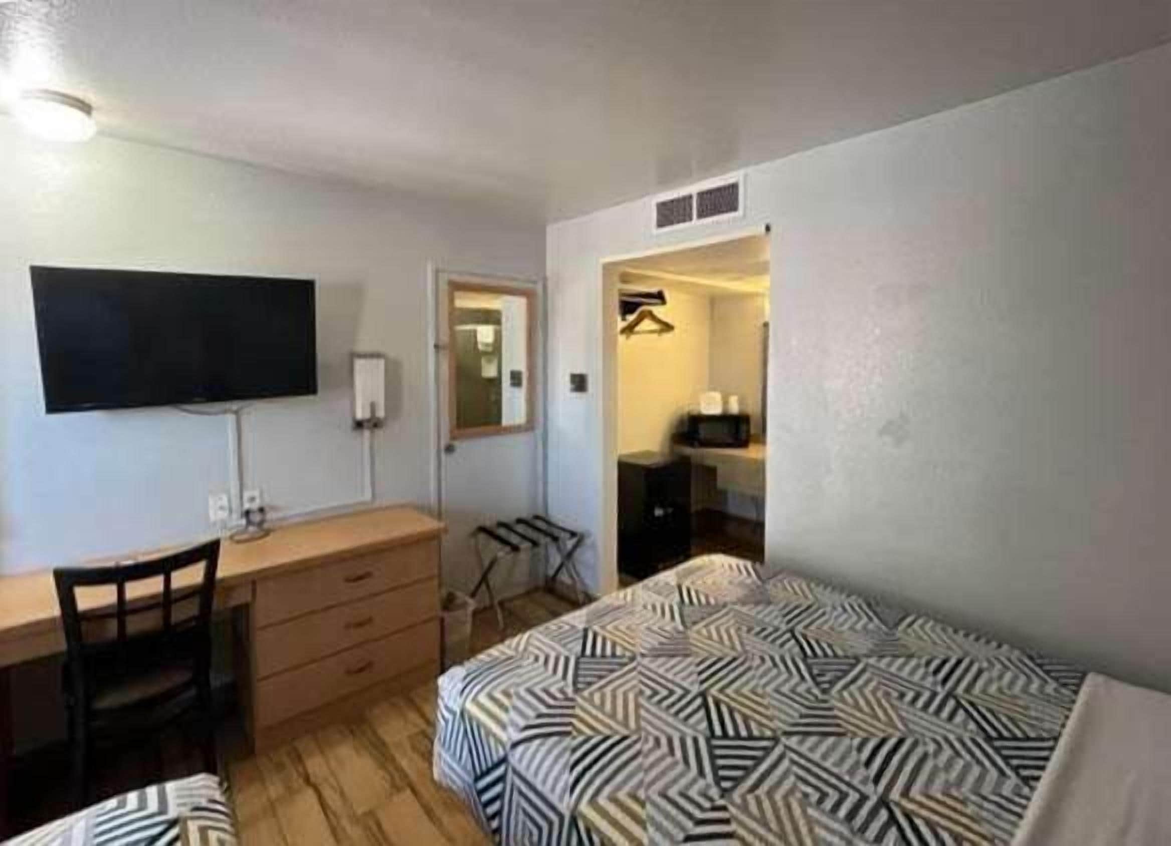 Habitación de hotel pequeña con cama, escritorio, silla, cómoda, televisor y baño visible a través de una puerta abierta.