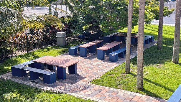 An outdoor patio area with multiple seating options, including benches and tables, surrounded by lush greenery and trees. - Motel 6 Riviera Beach, FL (West Palm Beach)