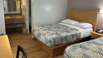 A hotel room with a bed, a bedside table with a phone, a wooden headboard, a mirror, a bathroom with a sink and toilet, and a chair.