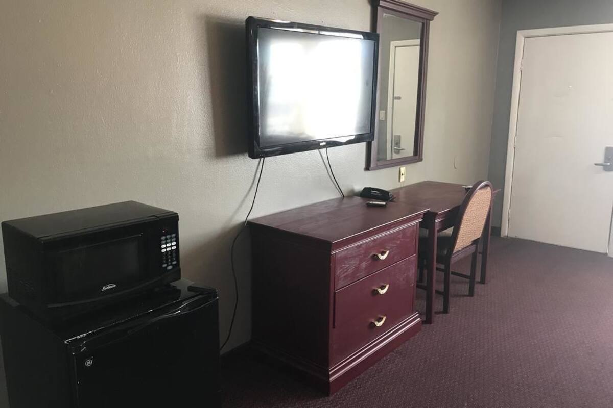 A room with a television mounted on the wall, a wooden dresser with drawers, a microwave on top of a refrigerator, and a chair.