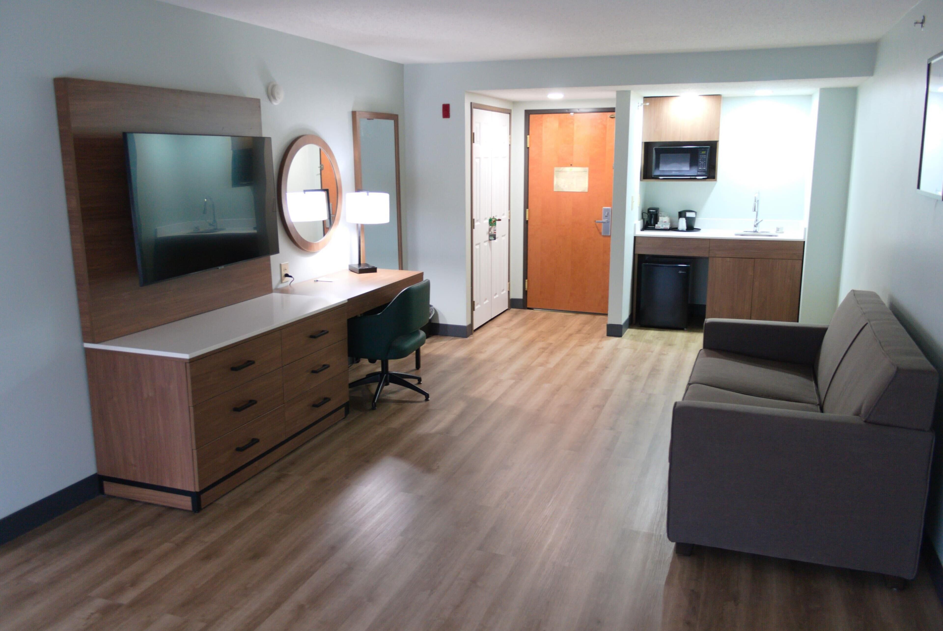 Una habitación de hotel moderna con piso de madera, televisor de pantalla plana, escritorio con lámpara, silla de oficina verde, sofá y kitchenette con microondas y cafetera.