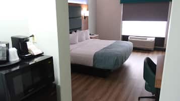 A hotel room with a bed, a desk with a chair, a microwave, and an air conditioner.