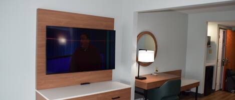 A hotel room with a TV mounted on the wall, a desk with a chair, a bedside table, and a mirror.