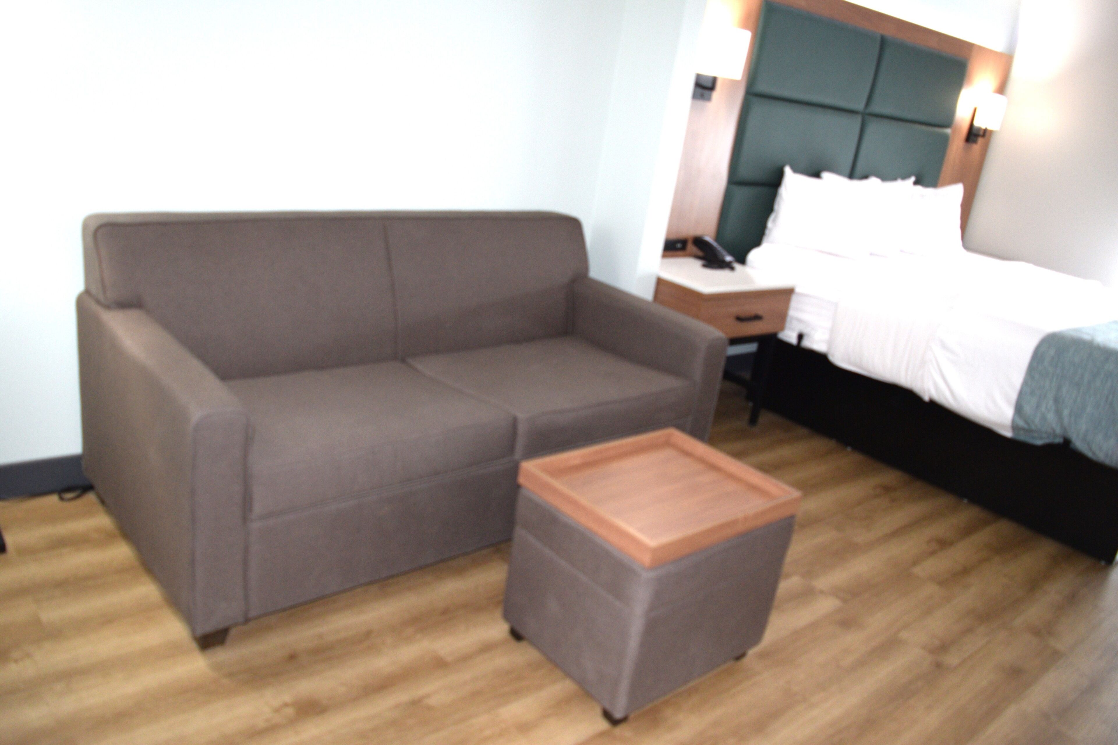 Habitación de hotel con sofá gris, una mesita de madera, cama con ropa blanca y una mesita de noche con lámpara.
