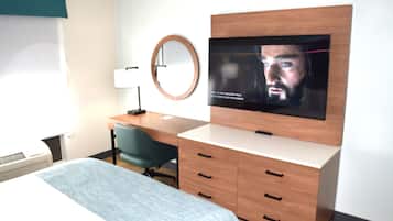 Una camera d'albergo con un letto, una scrivania, una sedia, una televisione, uno specchio e una finestra con tende.