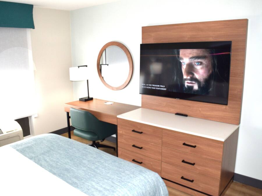 Una camera d'albergo con un letto, una scrivania, una sedia, una televisione, uno specchio e una finestra con tende.