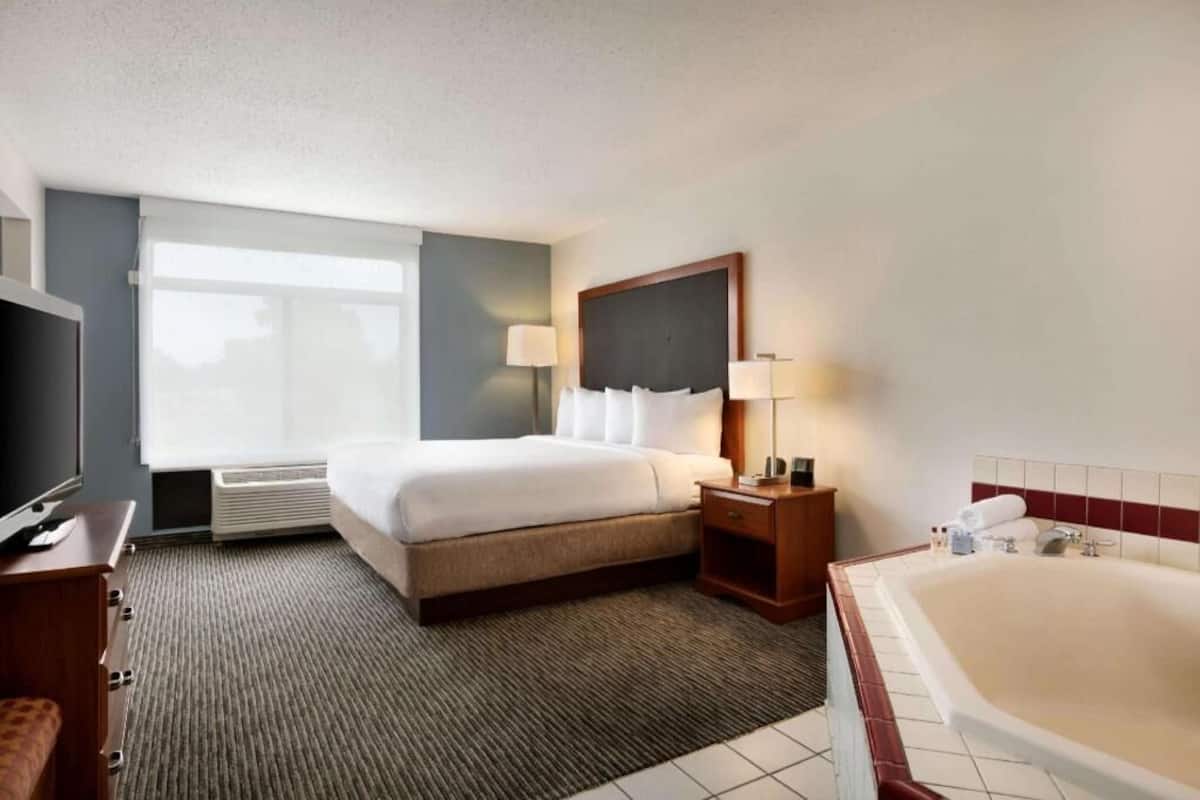 A hotel room with a bed, bedside table, lamp, TV, and a large bathtub.