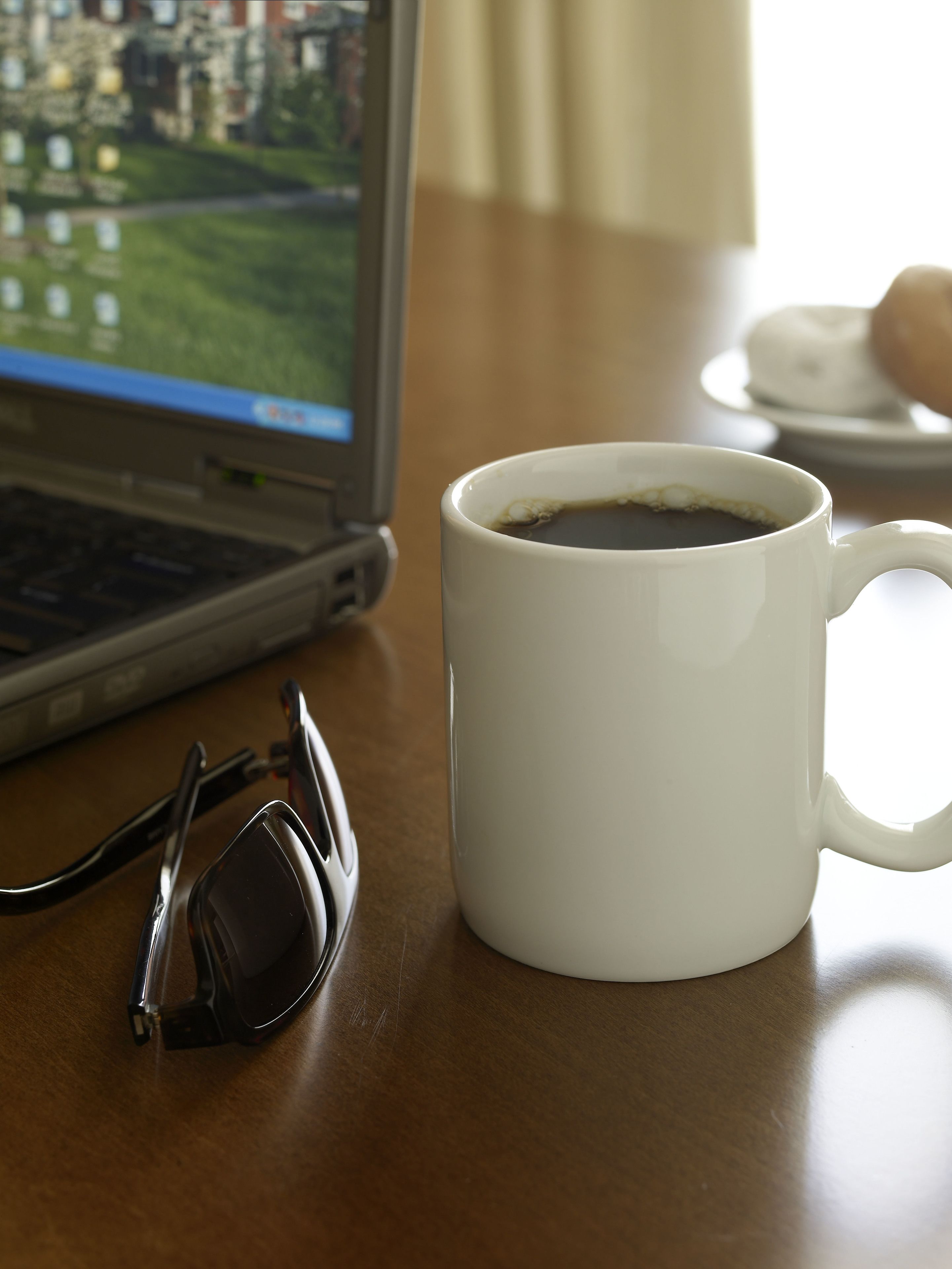Uma caneca branca com café sobre uma mesa de madeira, um par de óculos e um laptop exibindo uma paisagem.