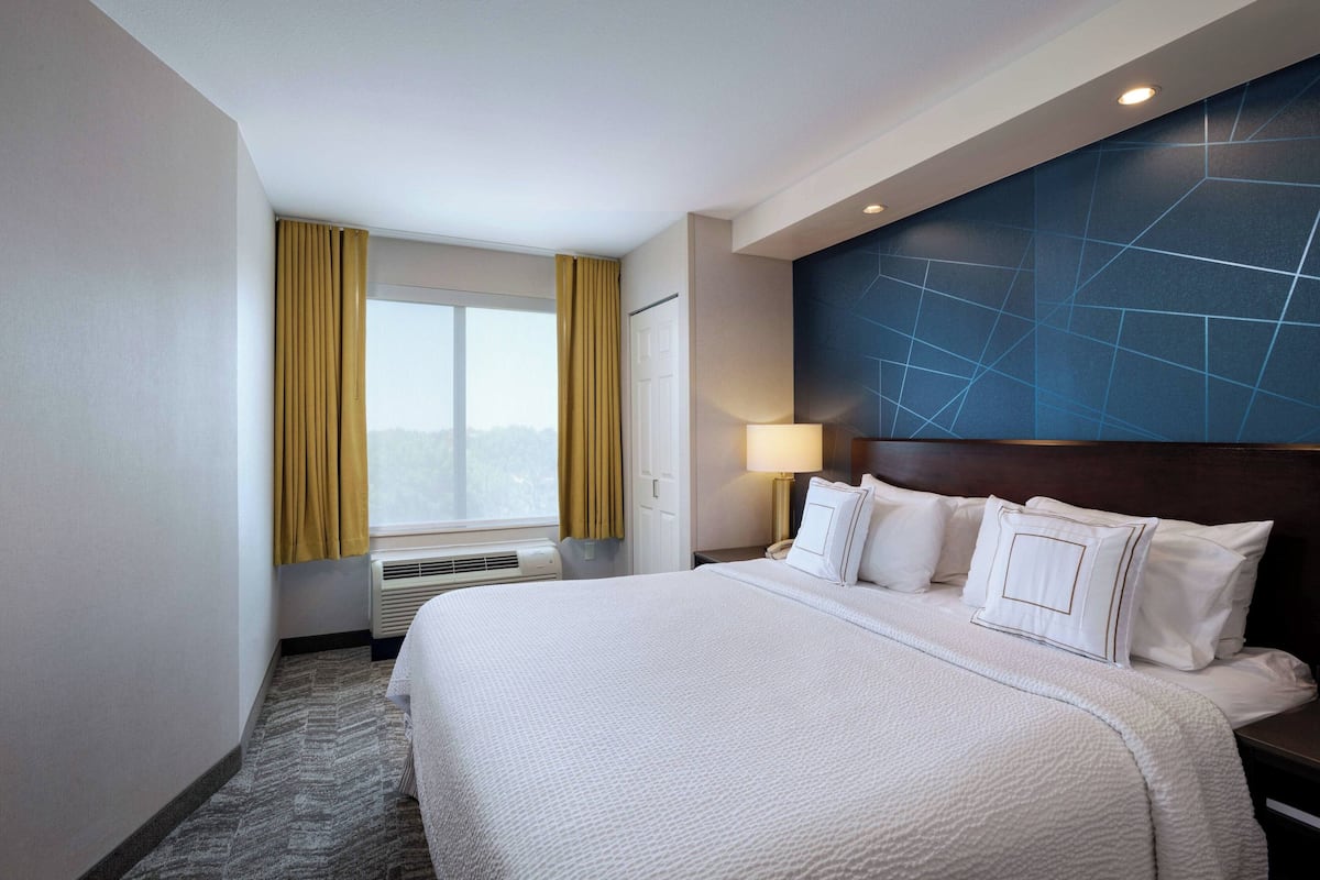 A hotel room with a large bed, a nightstand, a lamp, and a view of the outdoors through a window with curtains.