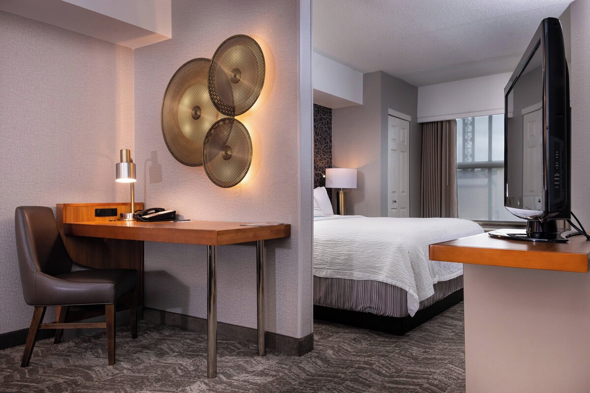 A hotel room with a desk, chair, bed, TV, and decorative wall lights.