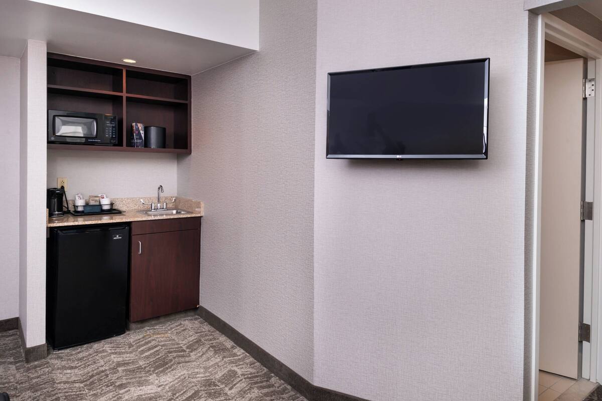 A compact hotel room with a kitchenette, a flat-screen TV mounted on the wall, and a patterned carpet.