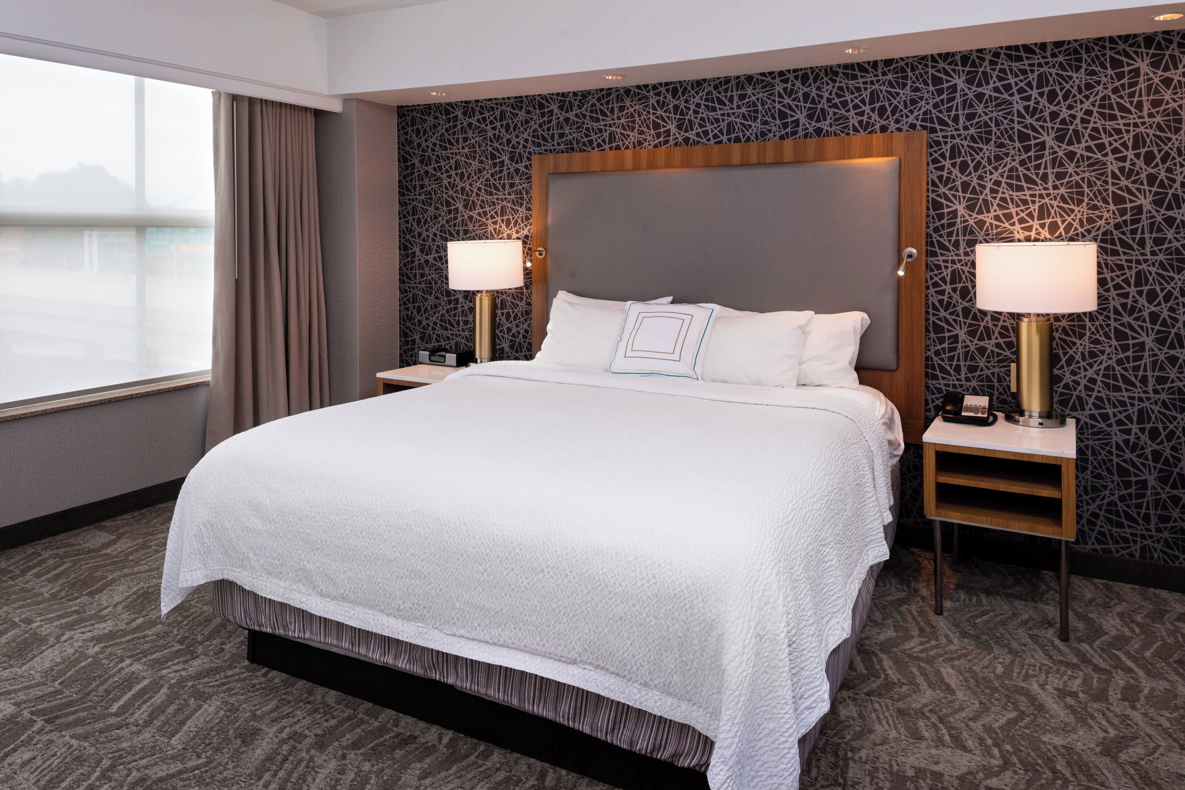 Habitación de hotel con una cama grande, dos mesitas de noche con lámparas, un papel tapiz decorado y vista a un cuerpo de agua a través del ventanal.