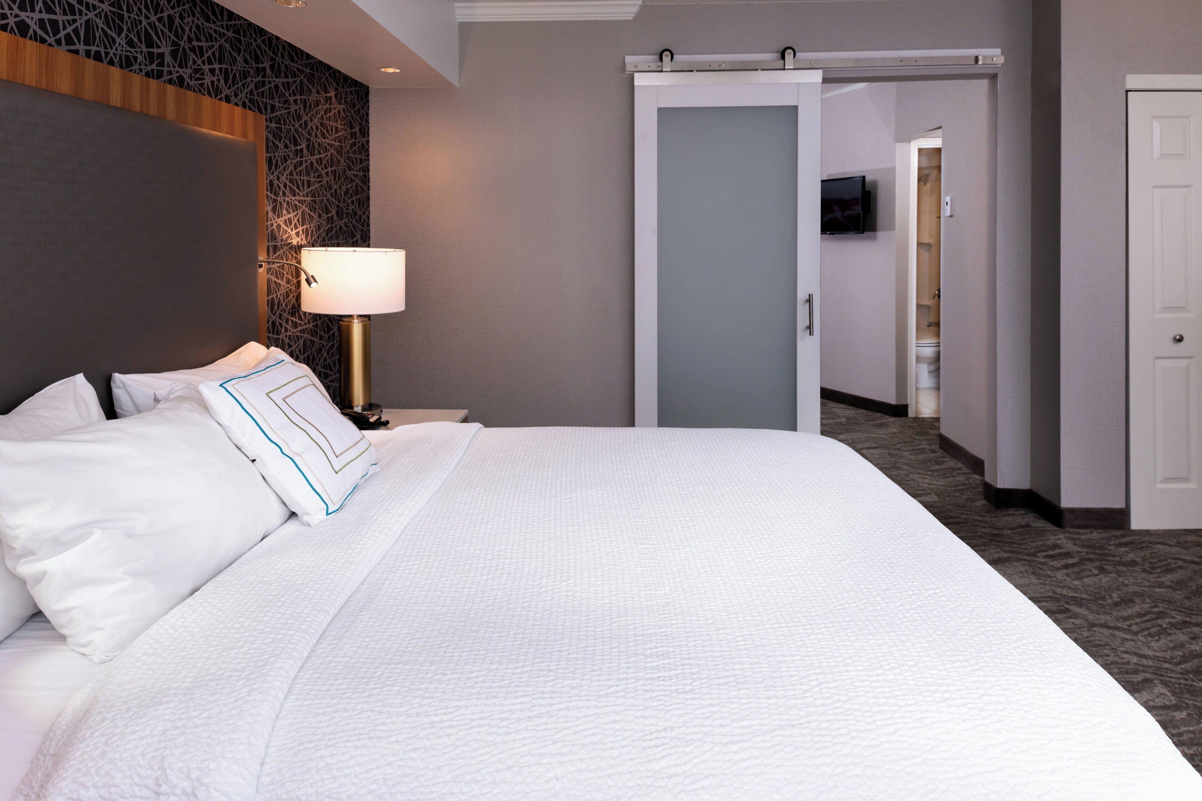 Una habitación de hotel con una cama grande, una lámpara de noche, un televisor en la pared y una puerta corredera que da al baño.