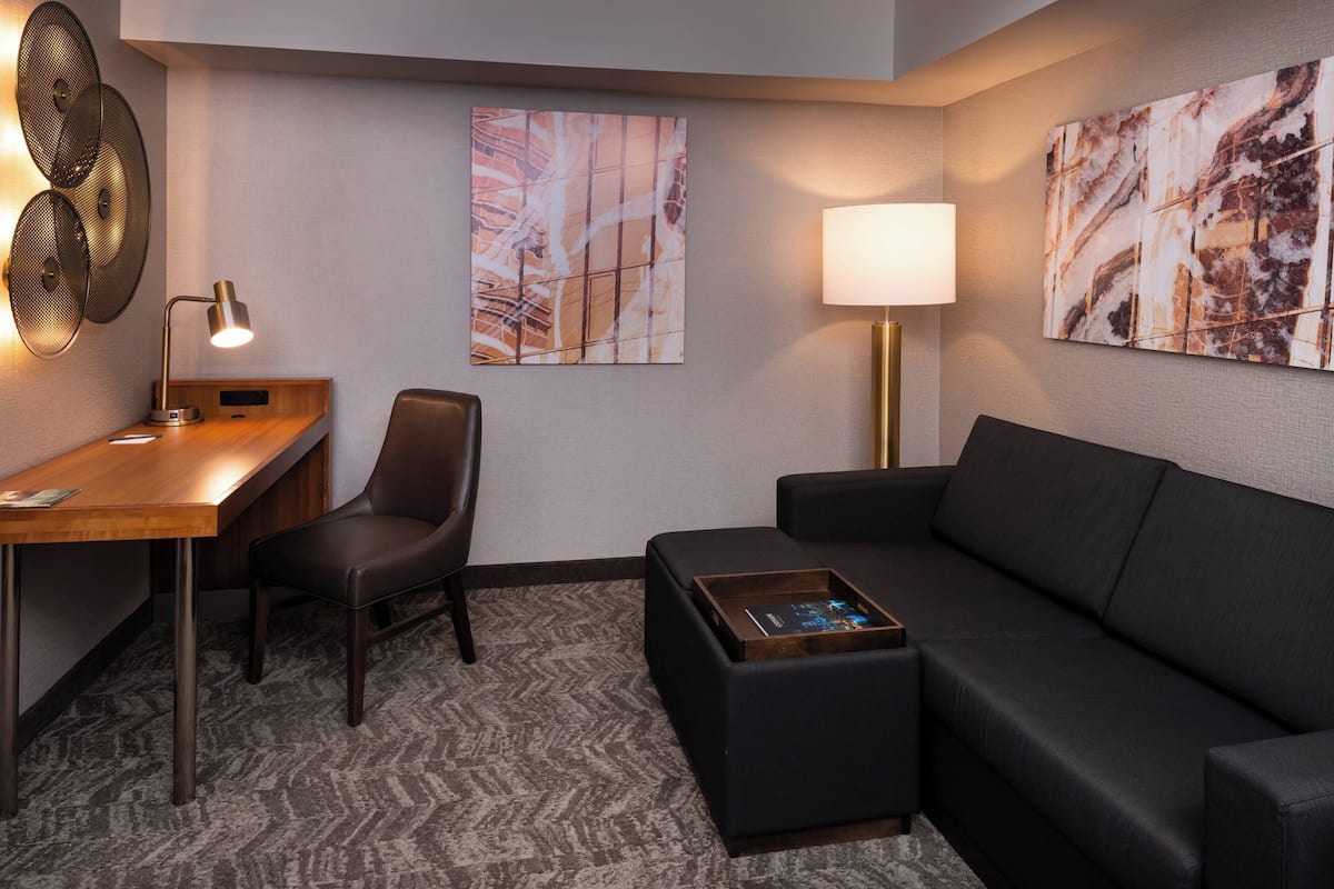A modern hotel room with a desk, a black leather chair, a black sofa, a standing lamp, and abstract wall art.