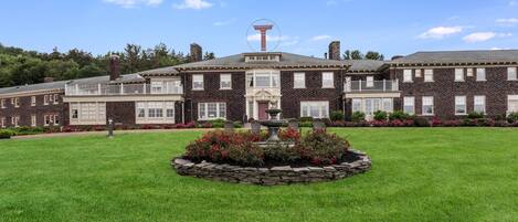 A large brick building with a central fountain, surrounded by a well-maintained lawn and flower beds.