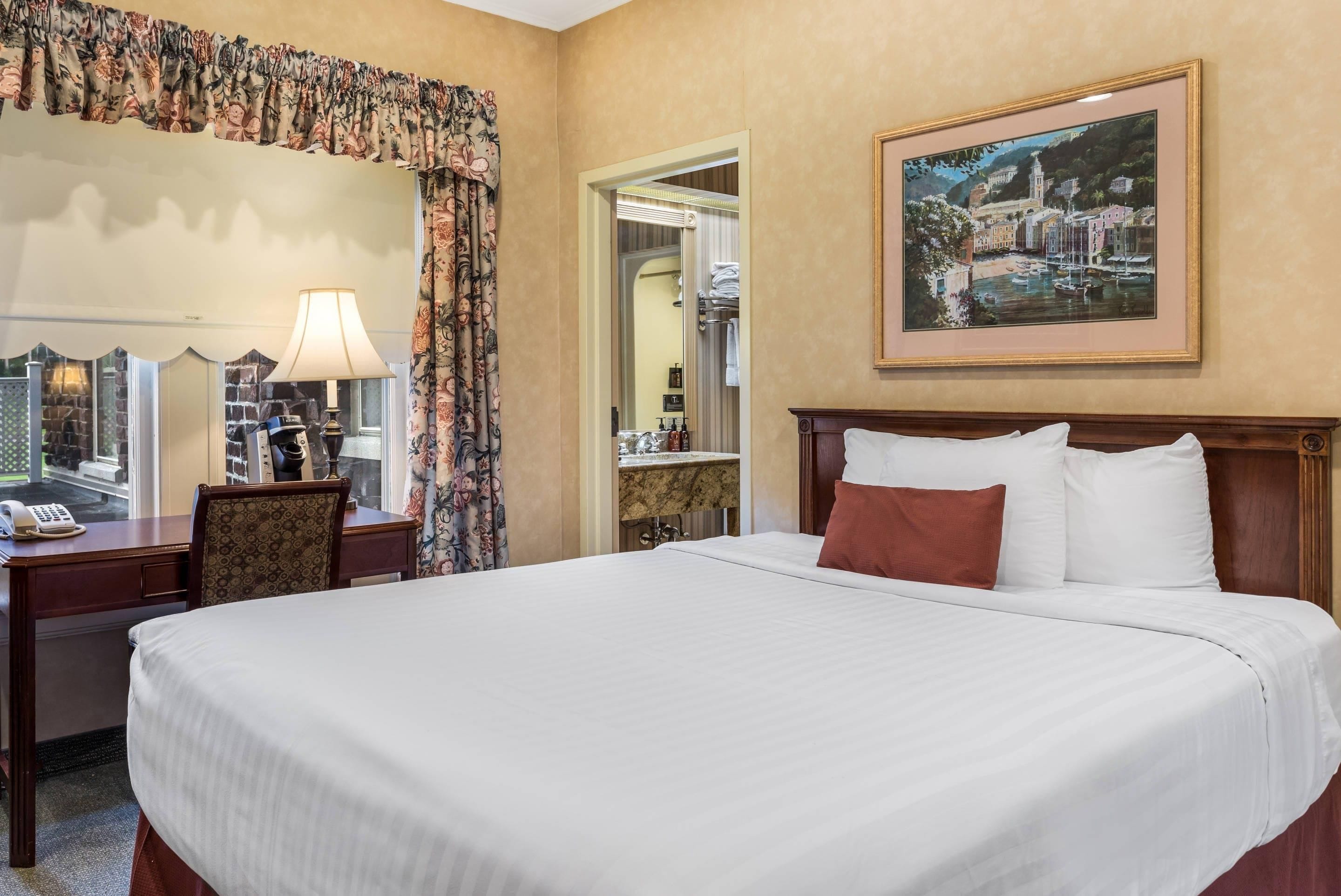 A hotel room with a large bed, a desk with a lamp, a chair, a bathroom mirror, and a framed painting on the wall.