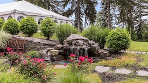 Una tienda de campaña junto a una cascada de piedra y un banco en un jardín con diversas plantas y árboles.