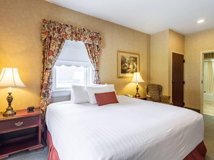 A hotel room with a large bed, two bedside lamps, a chair, a painting on the wall, and a door leading to a bathroom.