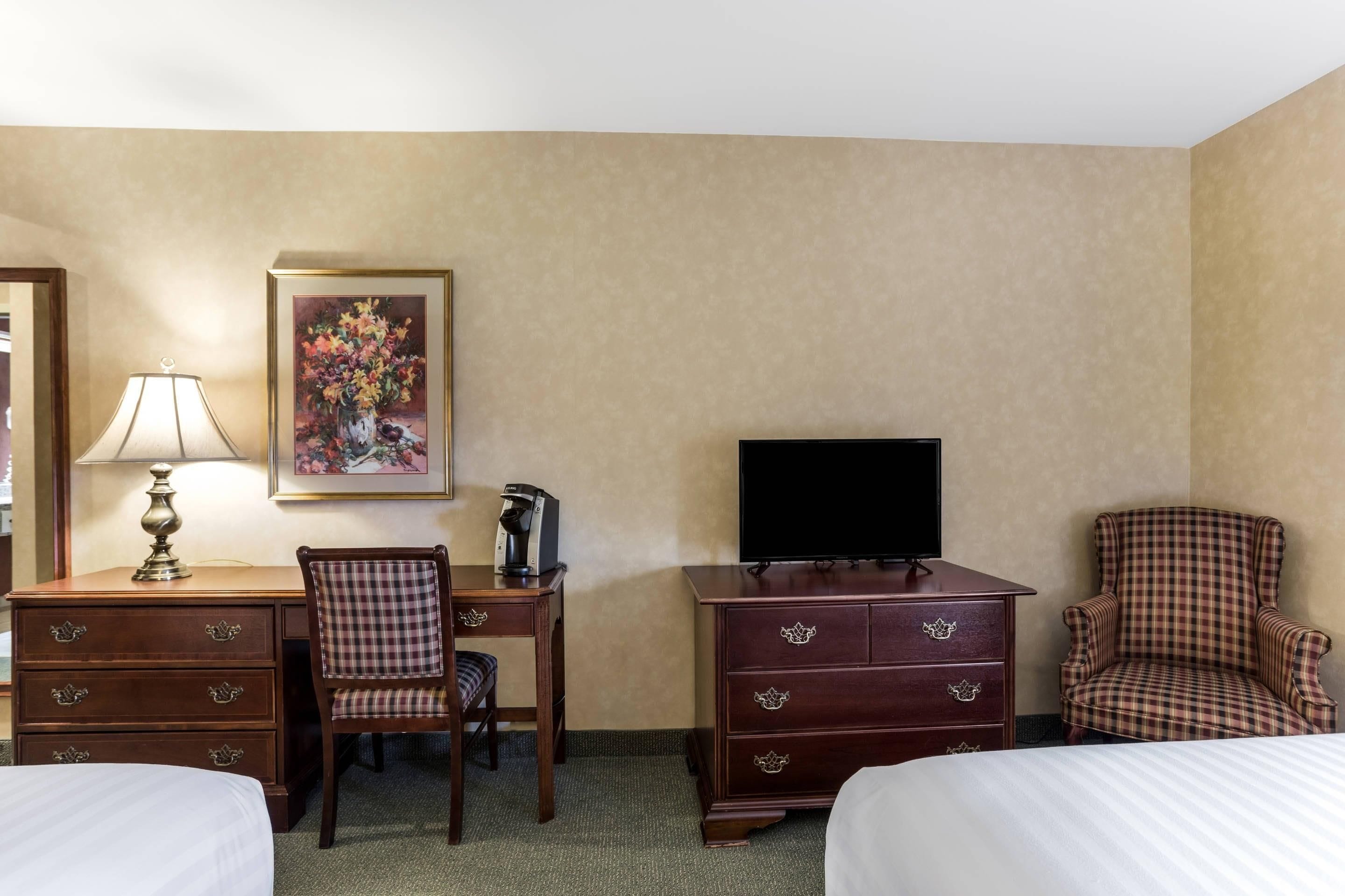 A hotel room with two beds, a desk with a lamp, a chair, a dresser, a television, and a painting on the wall.