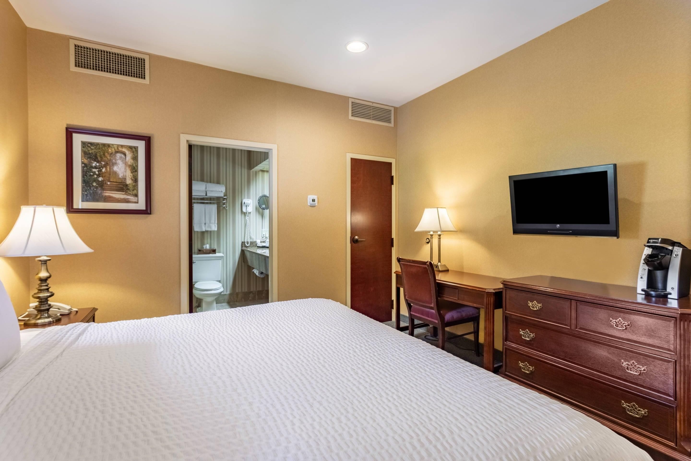 A hotel room with a bed, two bedside lamps, a television, a dresser, and a door leading to a bathroom.