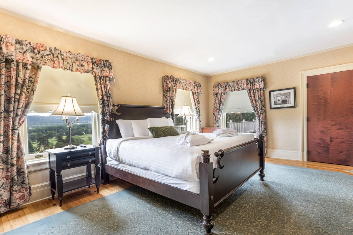 A bedroom with a four-poster bed, a nightstand, a lamp, and a view of the outdoors.
