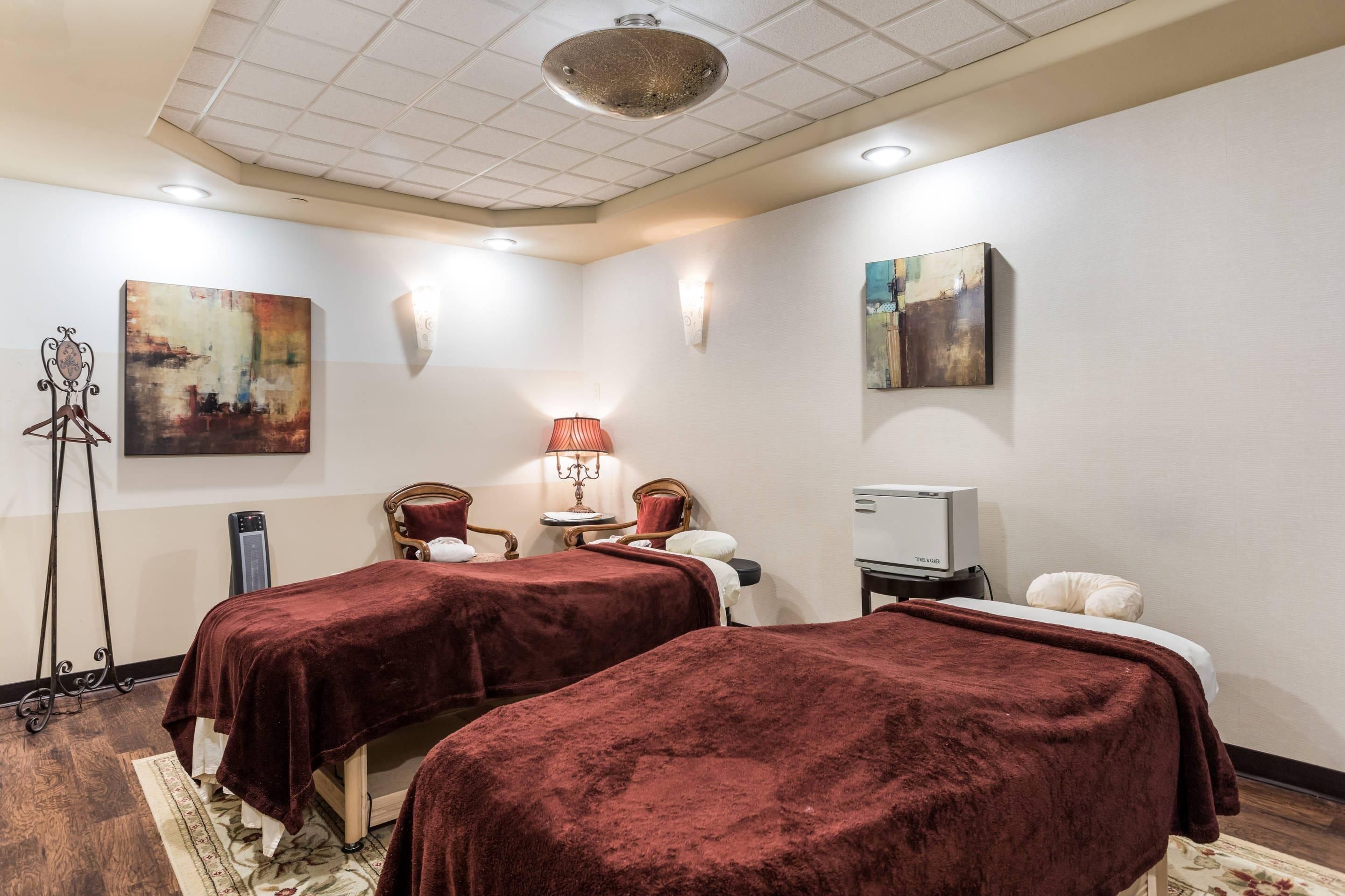 A room with two beds, a massage table, a massage chair, a small table with a lamp, and artwork on the walls.