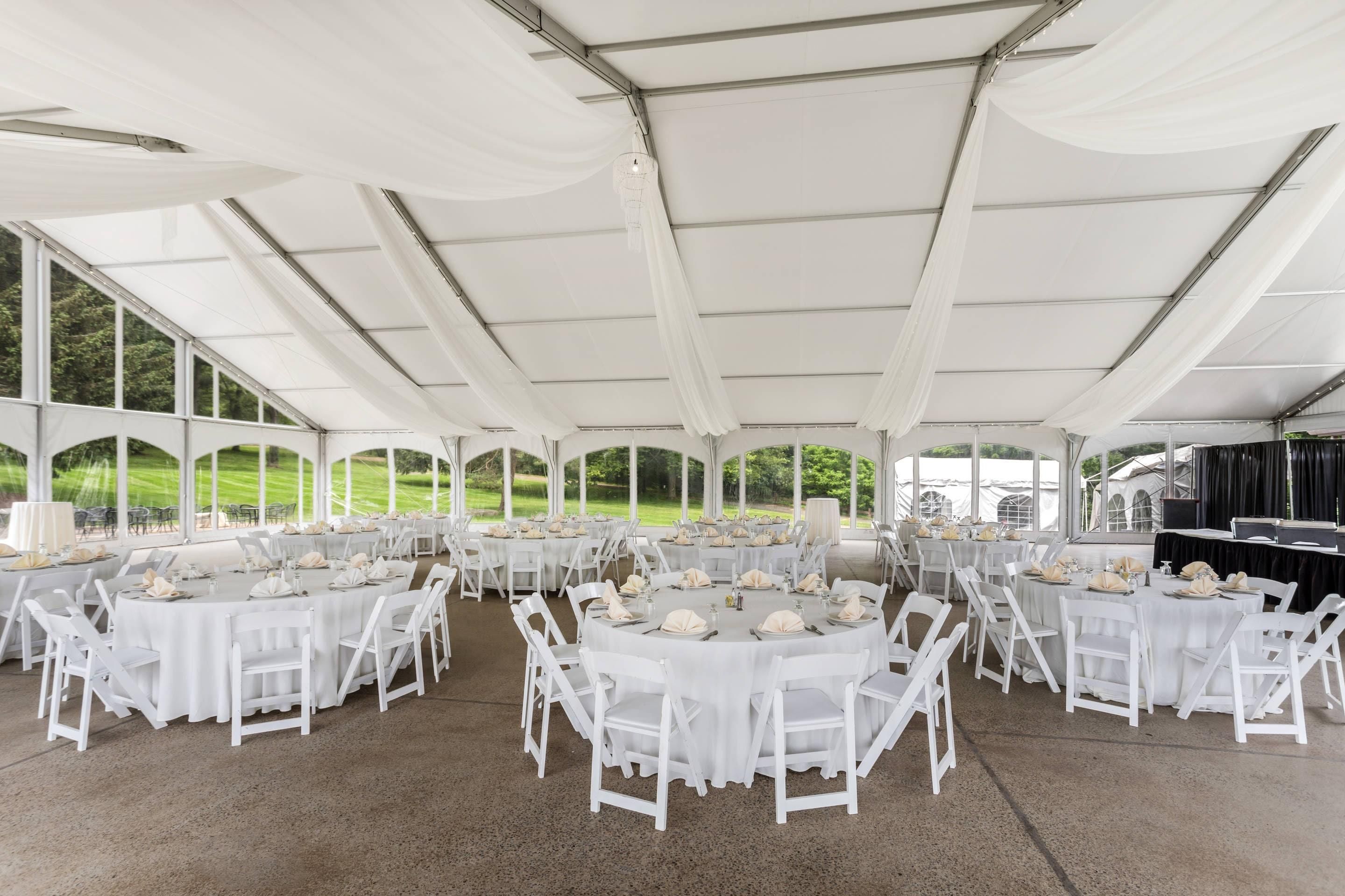 Un espace événementiel aménagé sous une tente, avec des tables rondes et des chaises blanches, de grandes fenêtres et une vue sur un terrain de golf.