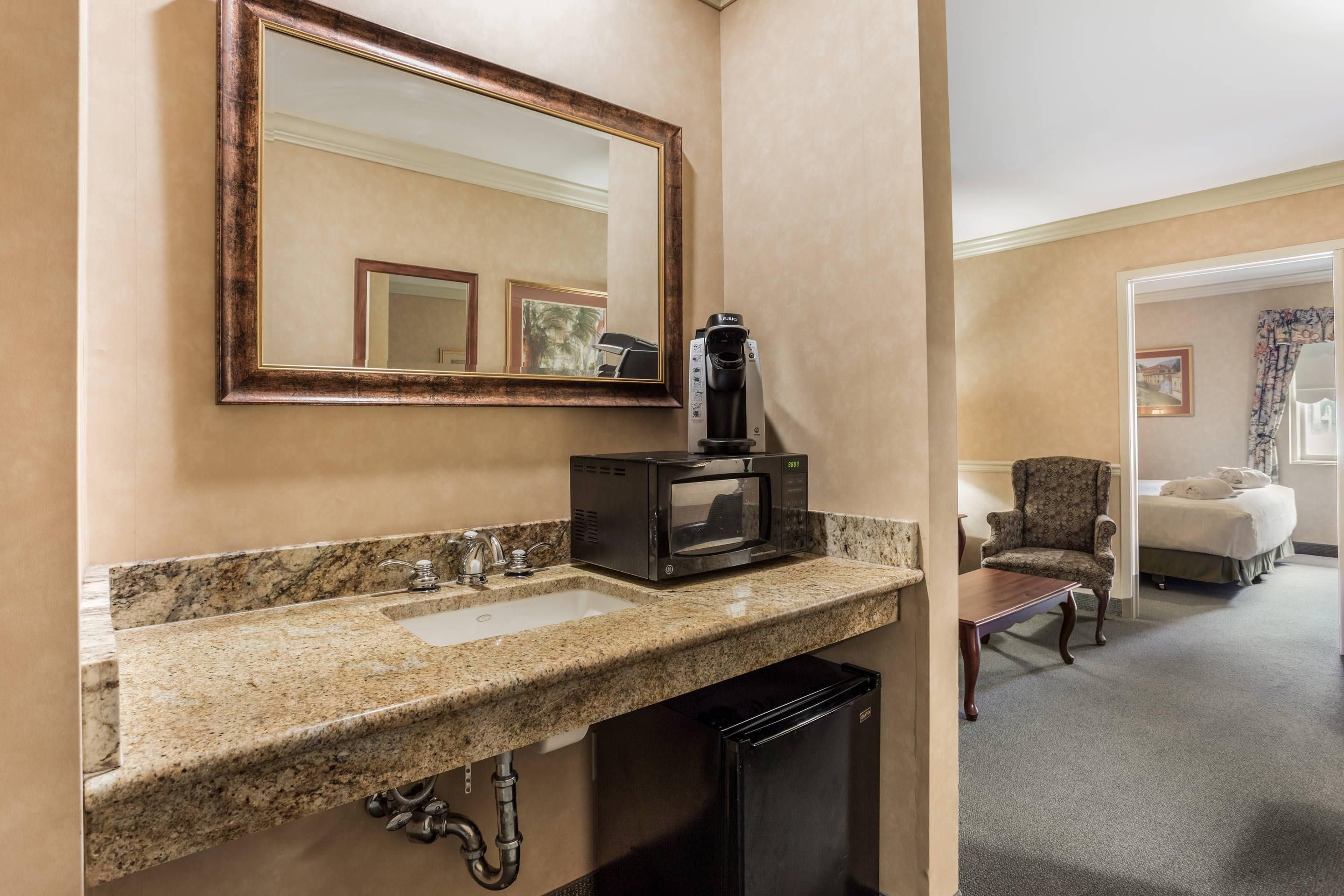 A hotel bathroom with a granite countertop sink, a mirror, a microwave, and a small black refrigerator.