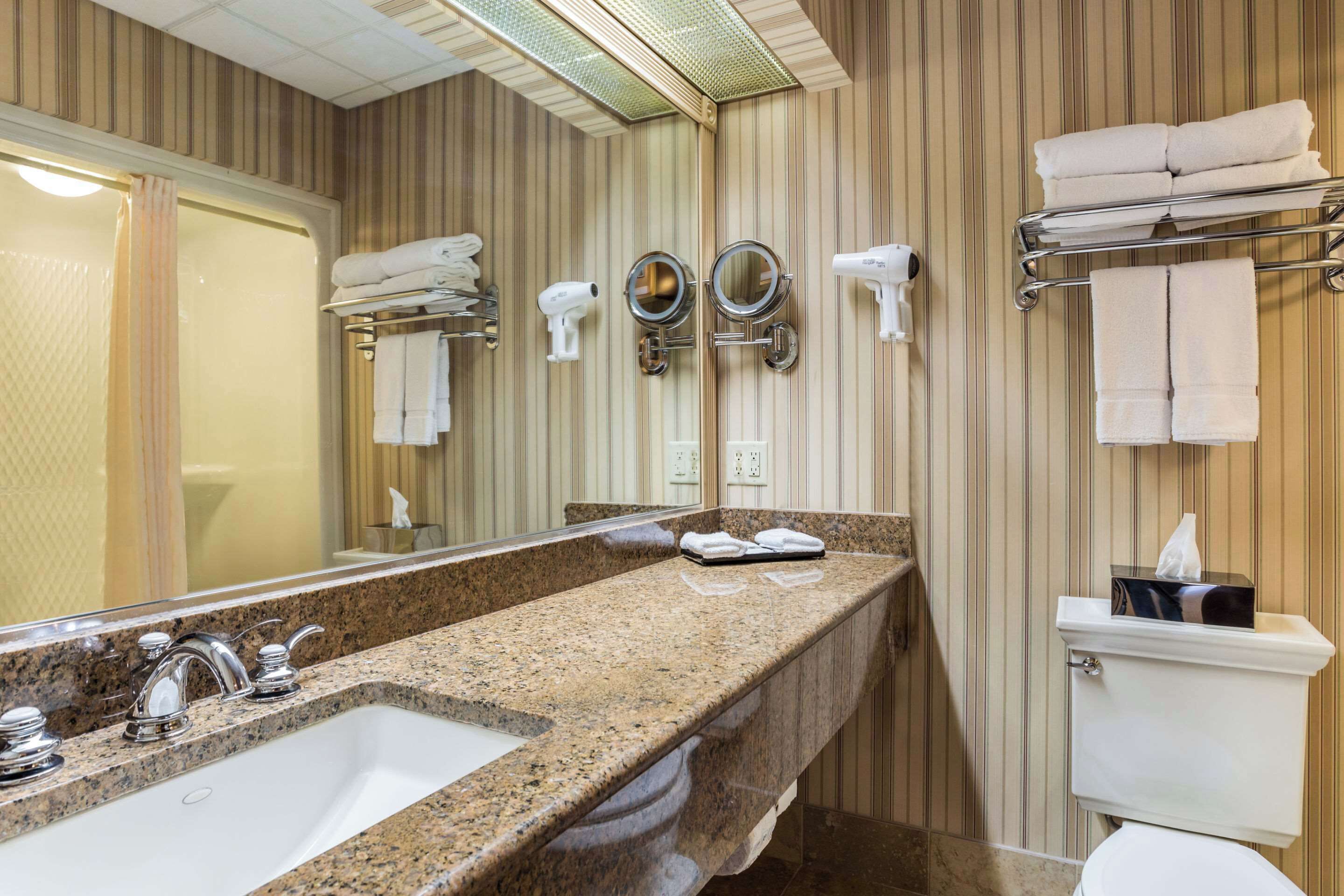Casa de banho com lavatório bancada de granito, duas espelhas, secador de cabelo e um porta-toalhas.