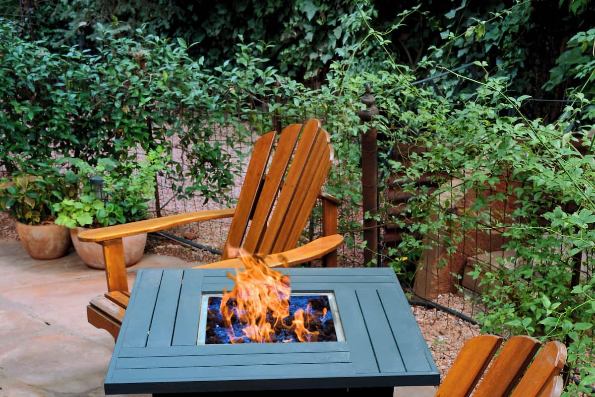 Una silla Adirondack de madera y una mesa cuadrada con una chimenea integrada, ubicadas en un patio de piedra rodeado de vegetación.