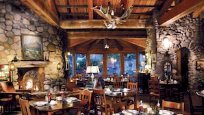 A rustic dining area with stone walls, wooden beams, and a chandelier. Tables are set with plates, cups, and cutlery.