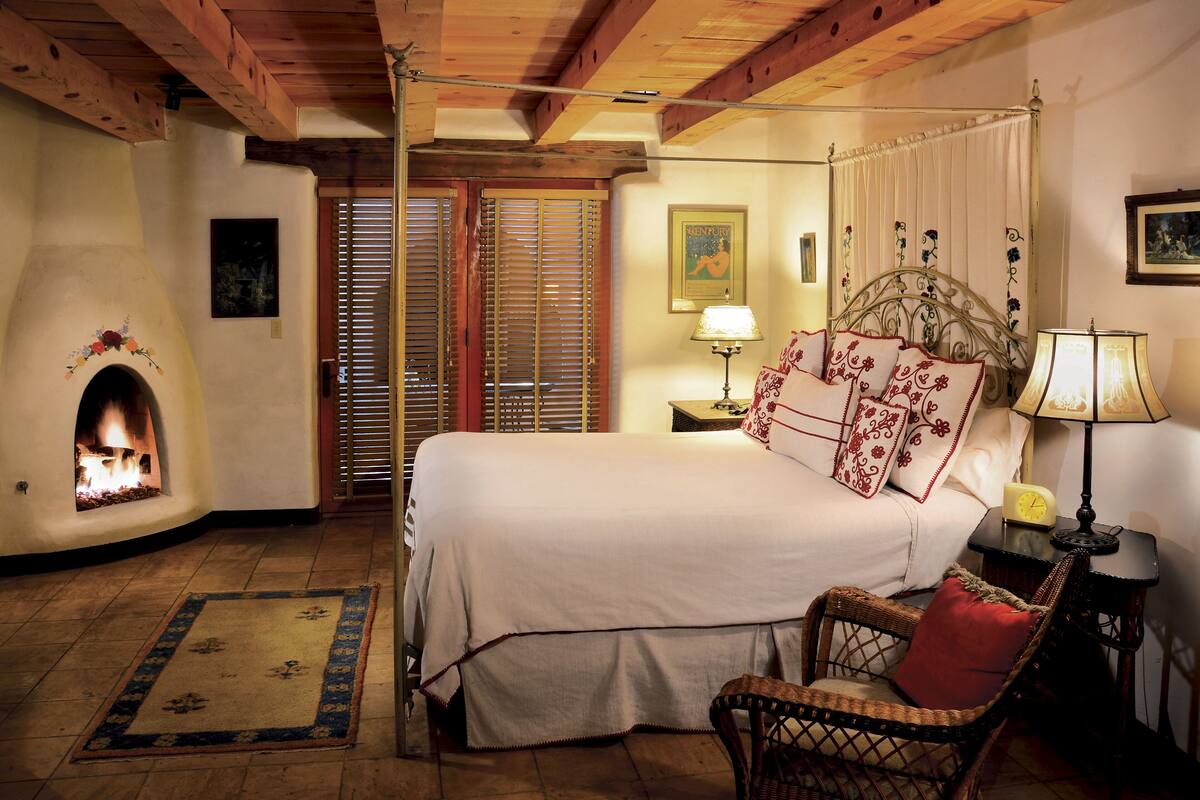 Un dormitorio con cama con dosel, chimenea, mesita de noche con lámpara y sillón con cojín rojo.