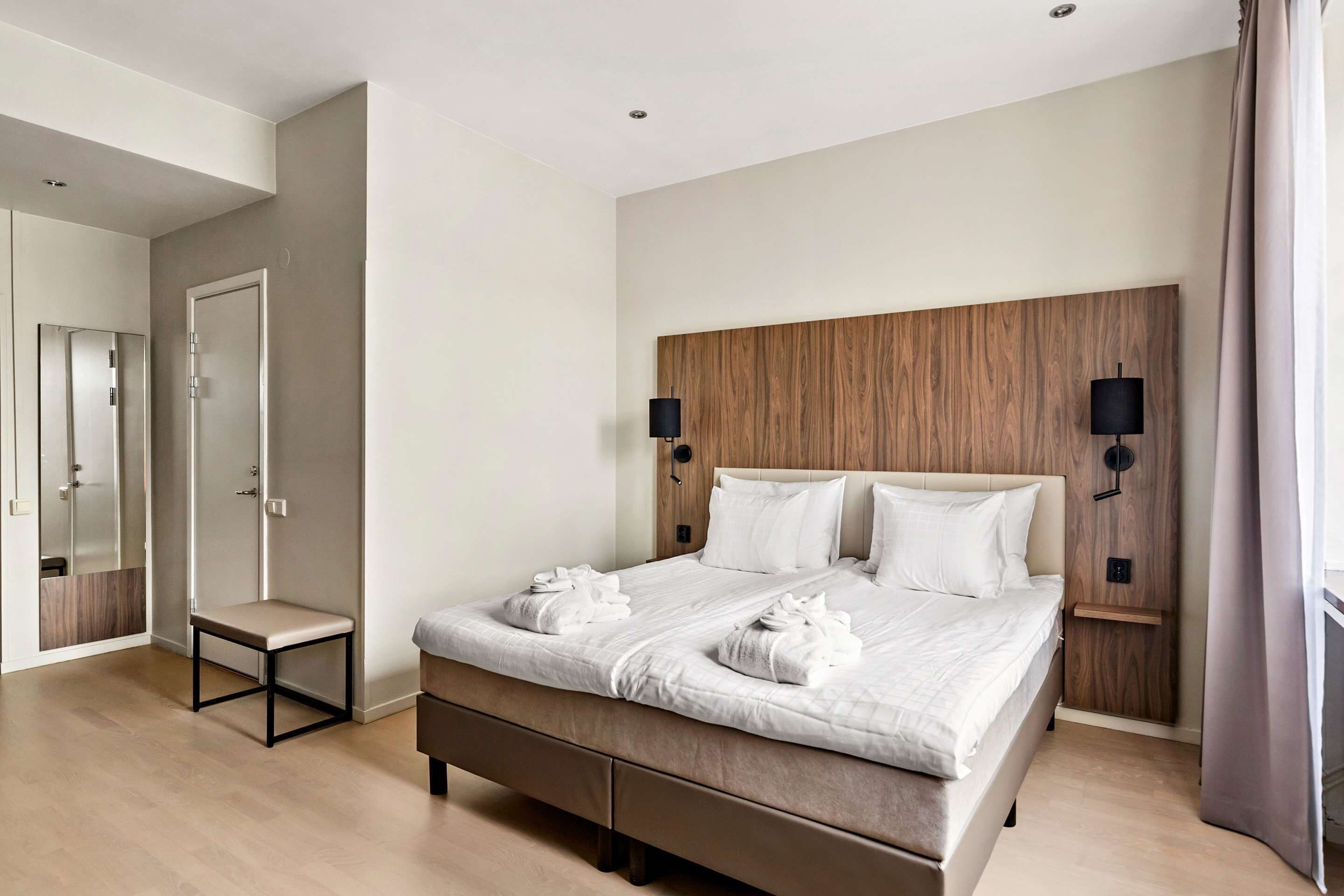 Camera d'albergo moderna con un letto grande, lampade sui comodini, una testiera in legno e un piccolo tavolino.